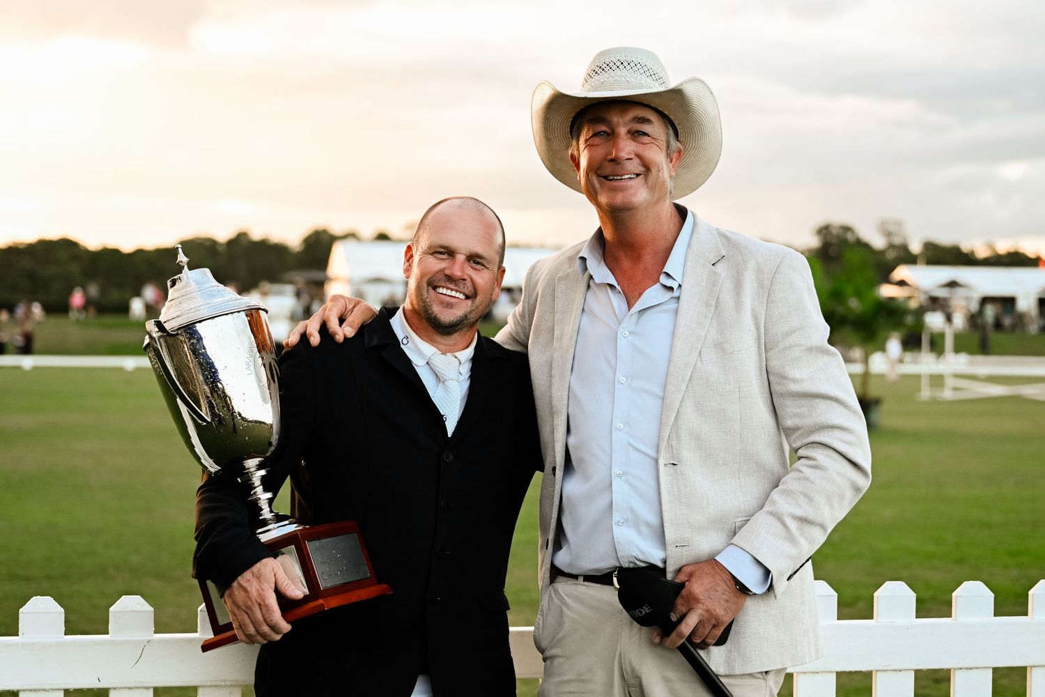 People celebrating at a polo club event with white picket fence in background.