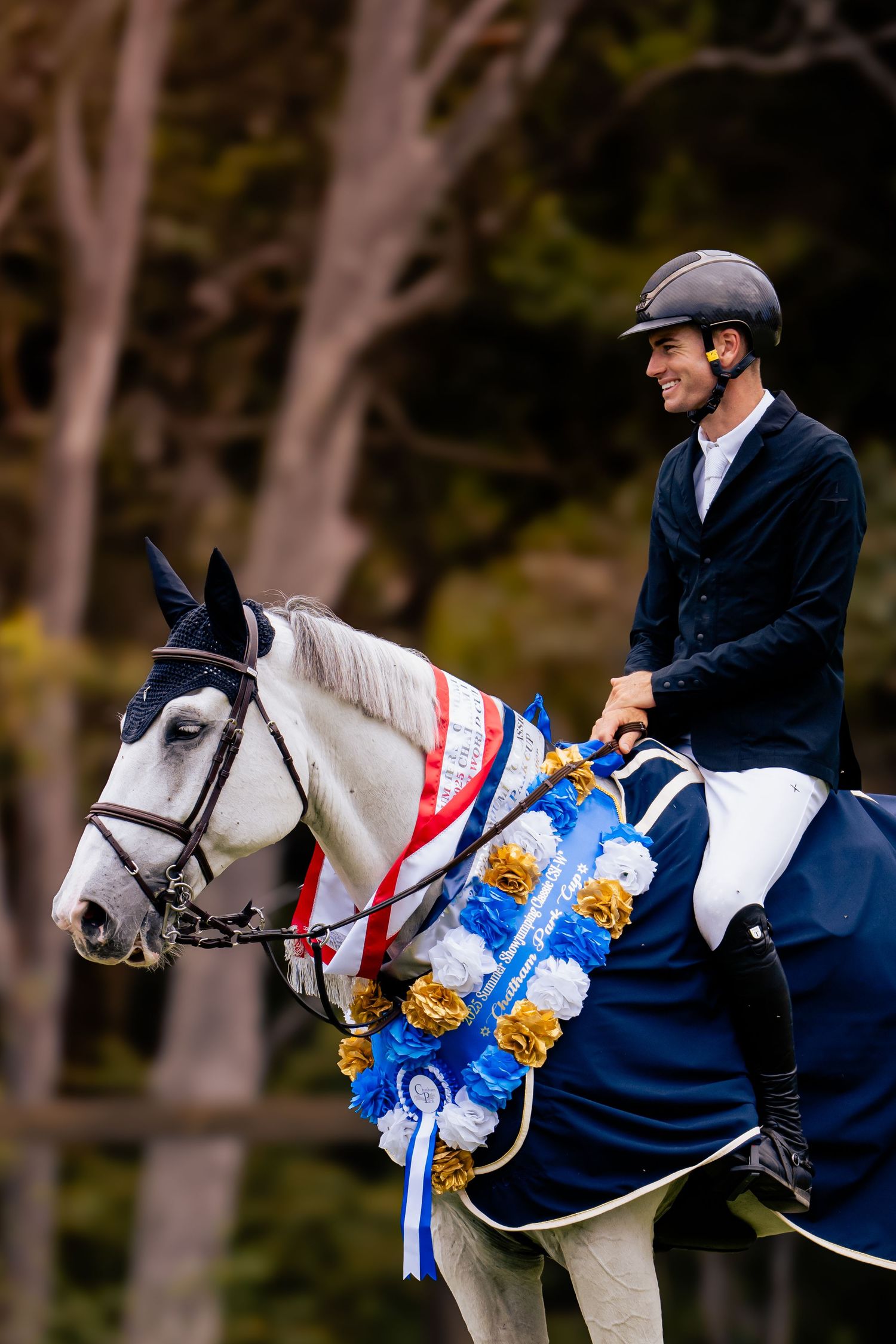 A rider in navy attire sits atop a decorated white horse with blue and red ribbons and awards at an equestrian event.
