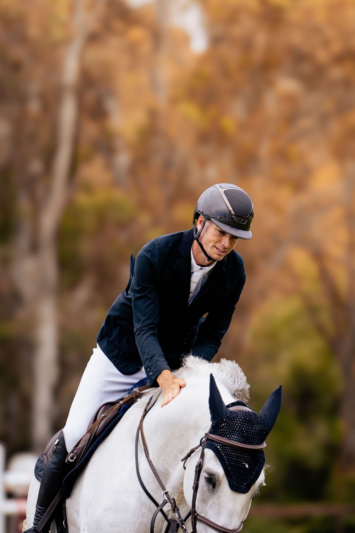 An equestrian rider in formal attire leans forward while riding a white horse with a navy bonnet against autumn foliage.