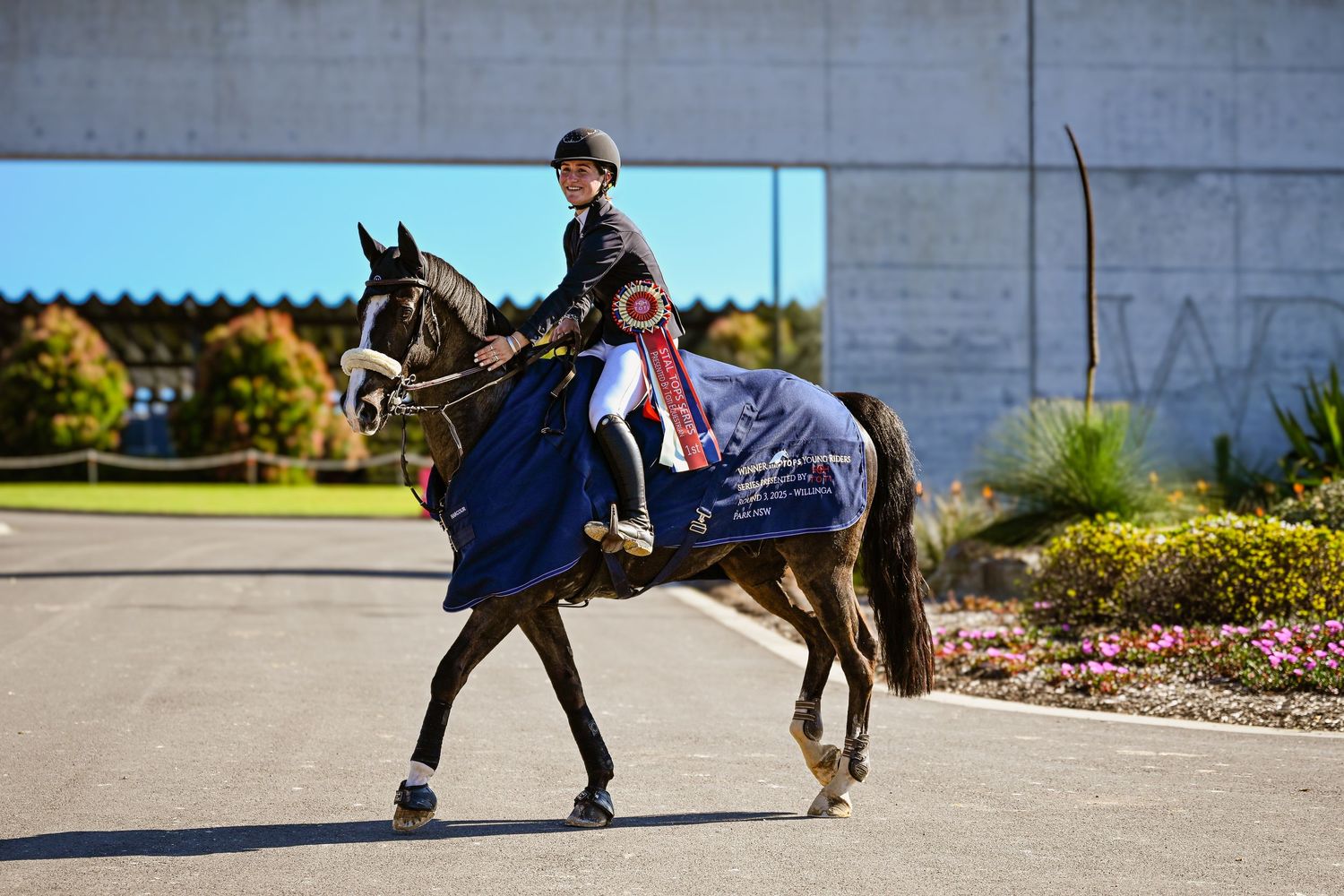 A young girl on a horse wearing a winning blue rug in front of a concrete archway.