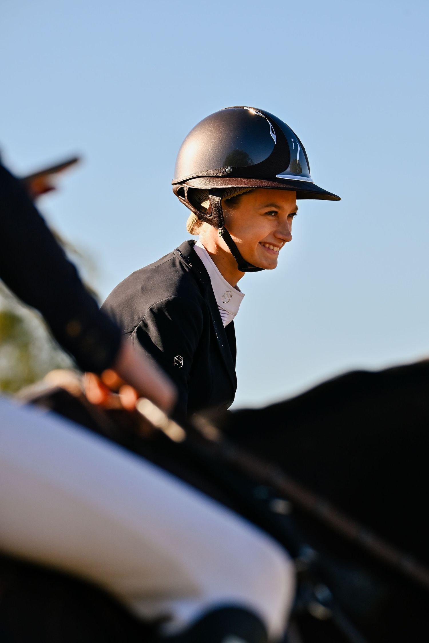 Equestrian rider in black jacket and helmet smiling against blue sky during competition.