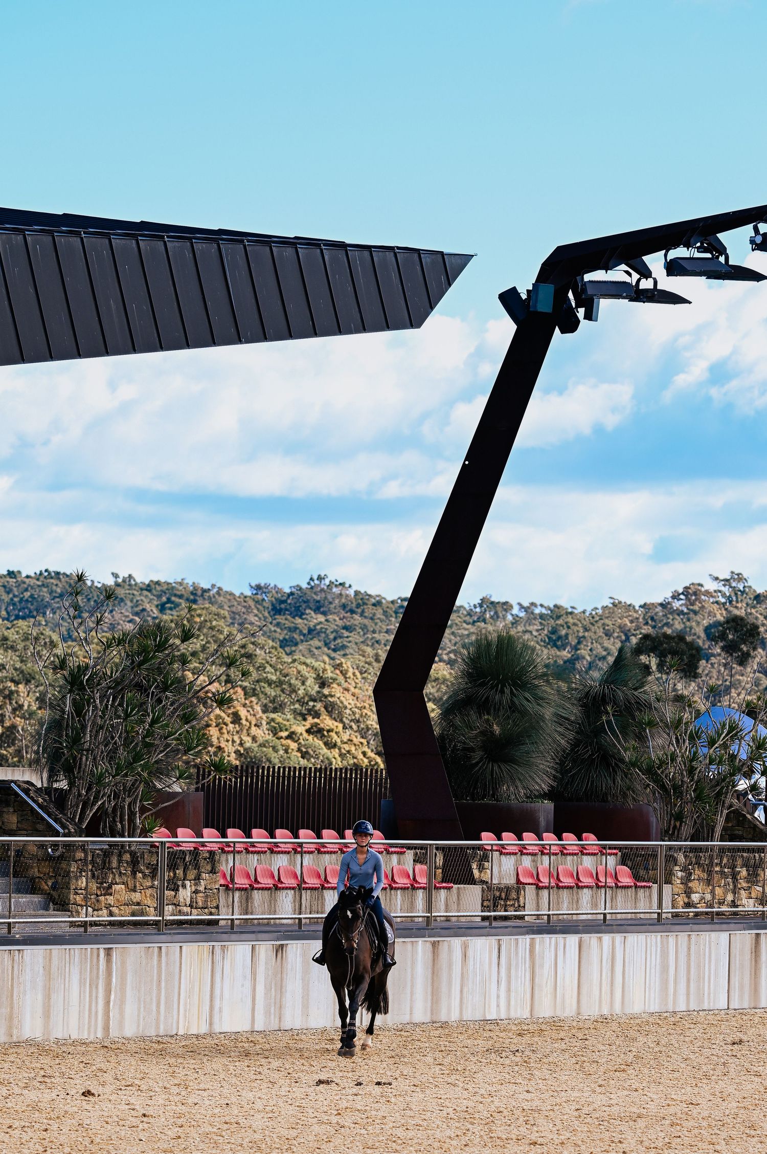 A rider on horseback practices in an outdoor arena with red stadium seats and a modern architectural structure overhead.