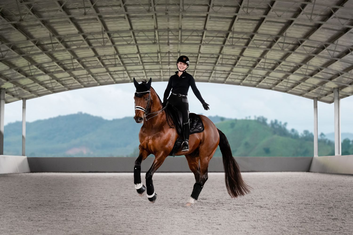 Covered indoor arena with mountain views where a horse and rider are preforming a high level dressage movement with one hand and smiling.