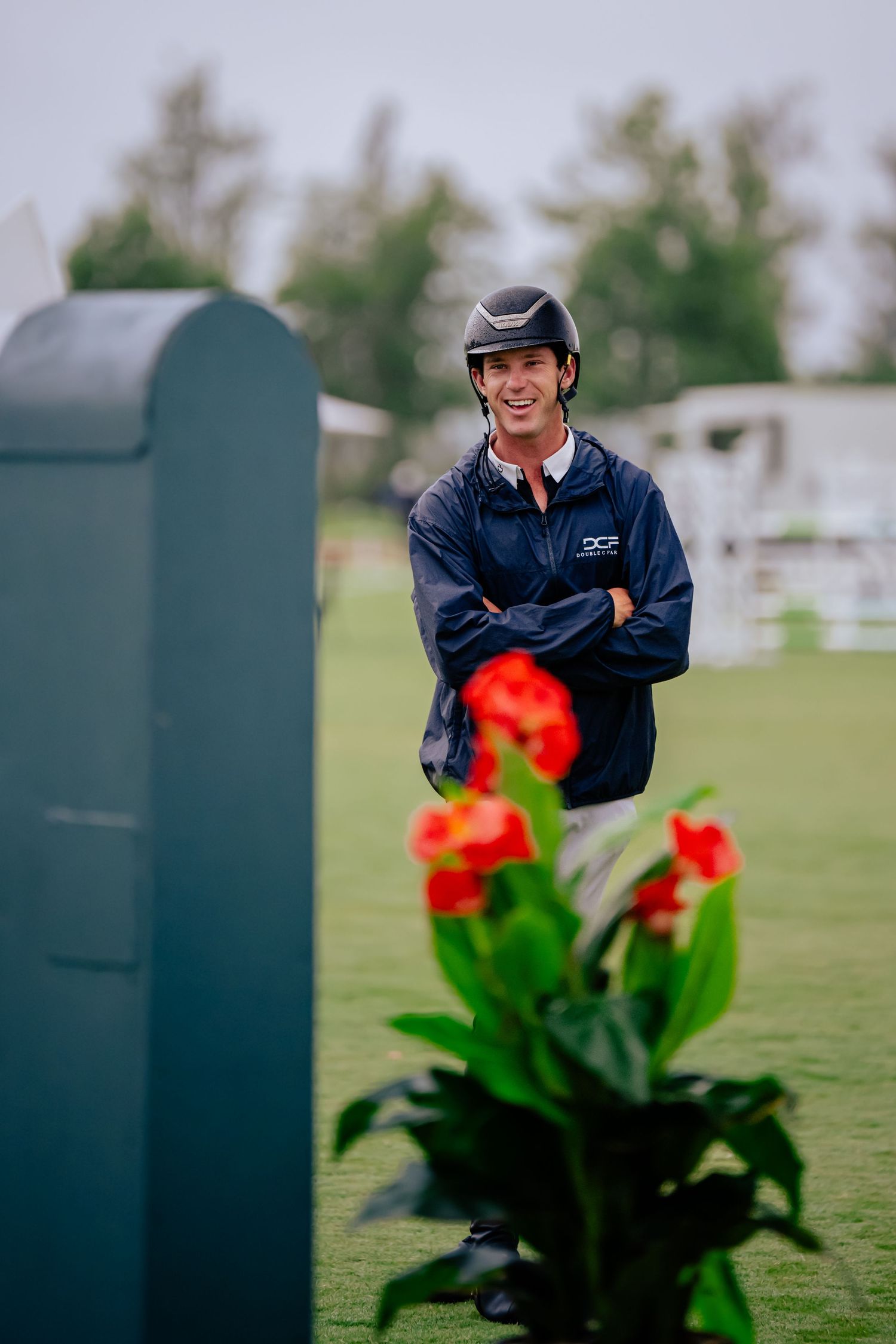 A person in a navy jacket and riding helmet stands behind bright red flowers.