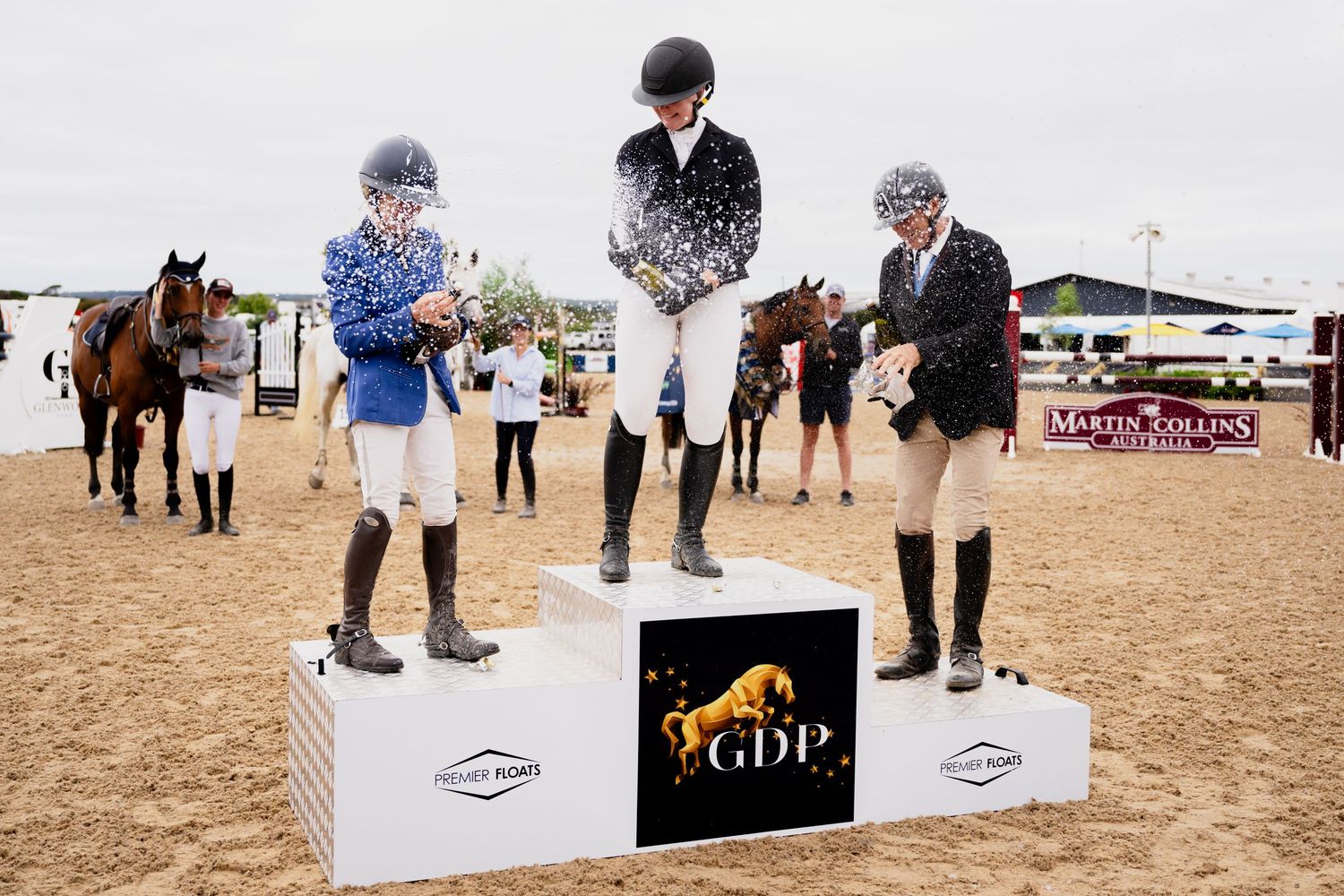Show jumping competitors prepare at arena entrance with numbered gates and banners.