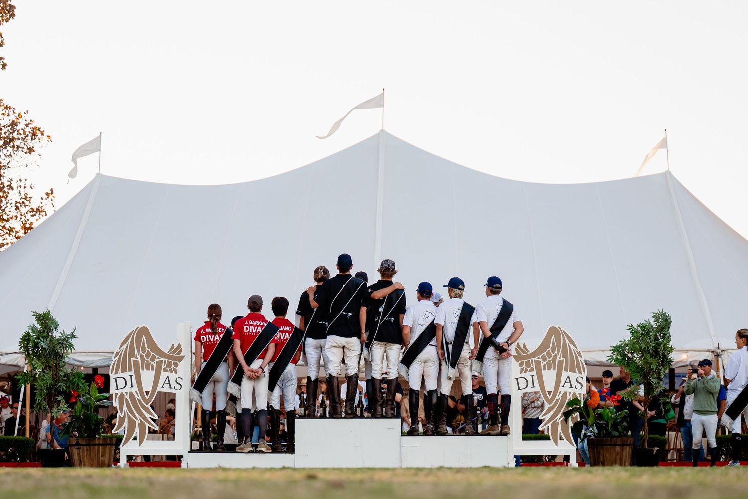 Polo teams stand on a podium before a white tent at an outdoor awards ceremony during a sunny event.