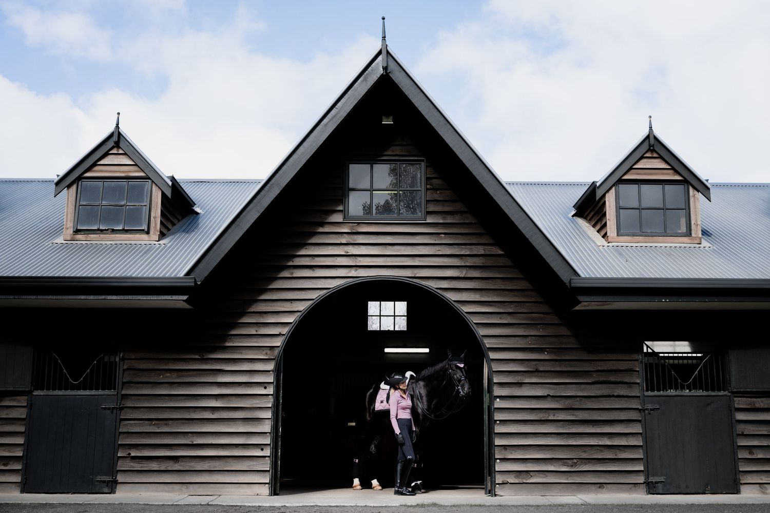 Row of black wooden stables with dormers under cloudy sky at equestrian facility.