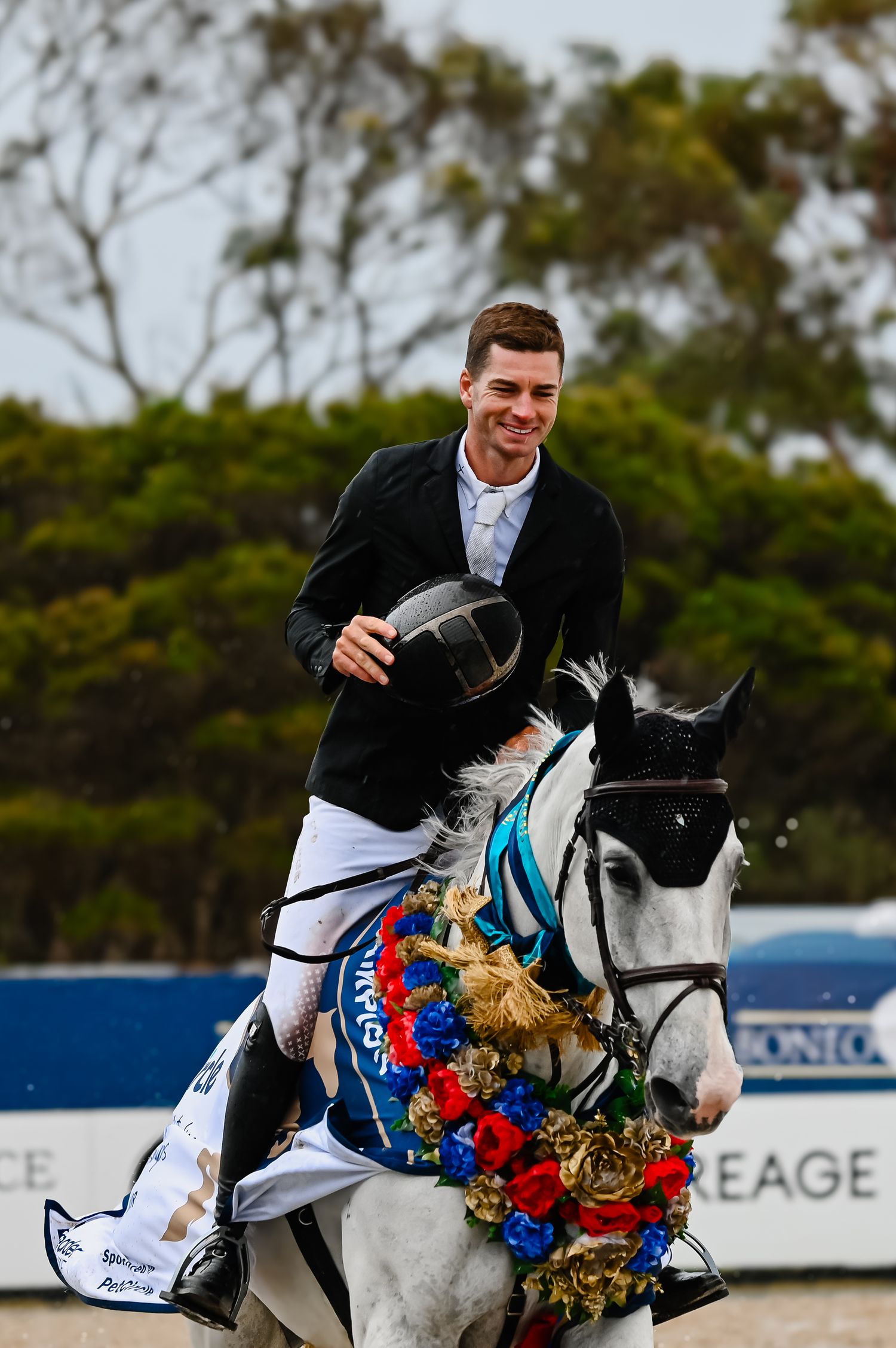 An equestrian in formal attire celebrates atop a decorated white horse wearing a blue and red garland at a competition.