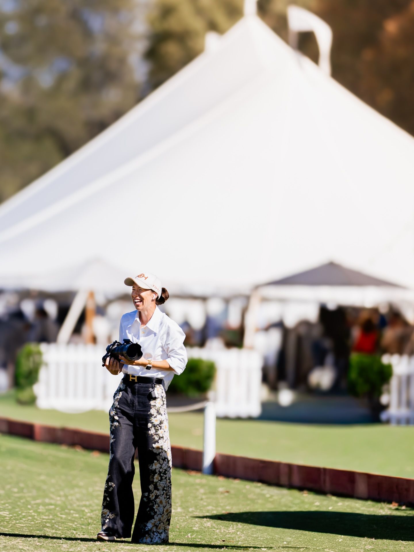 Photographer in white shirt and patterned pants stands on green lawn near white event tent at outdoor polo match.