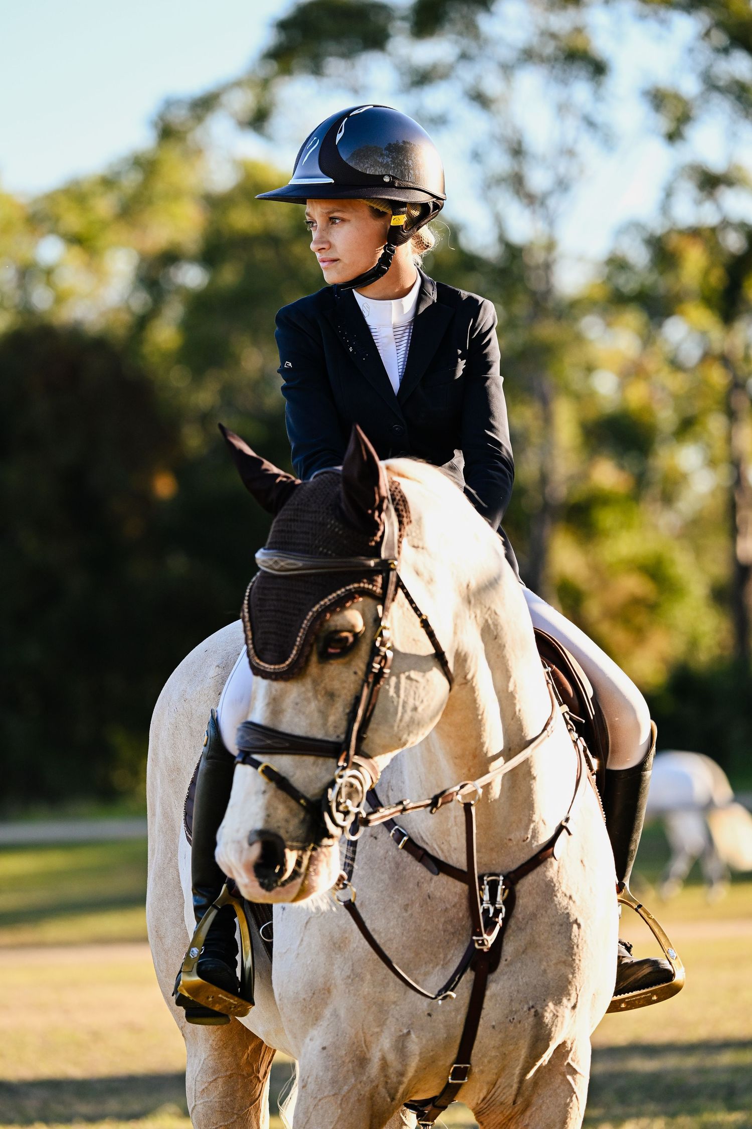 Equestrian in navy jacket riding white horse during competition in sunny outdoor setting.