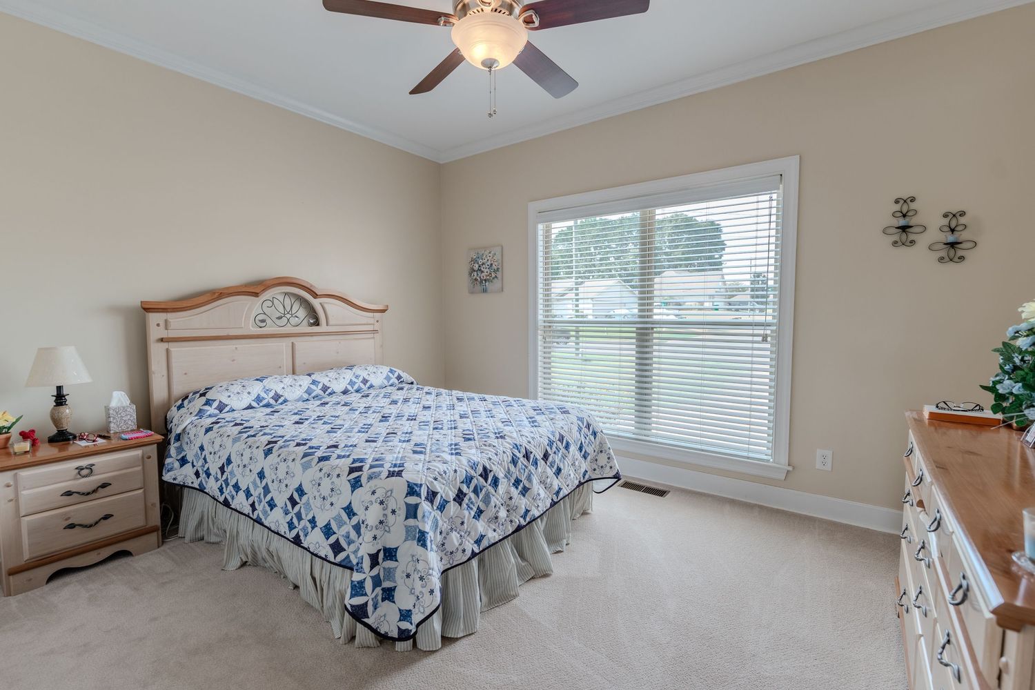 Cozy bedroom with blue and white floral bedding, light beige walls, window with natural light, and wooden furniture.
