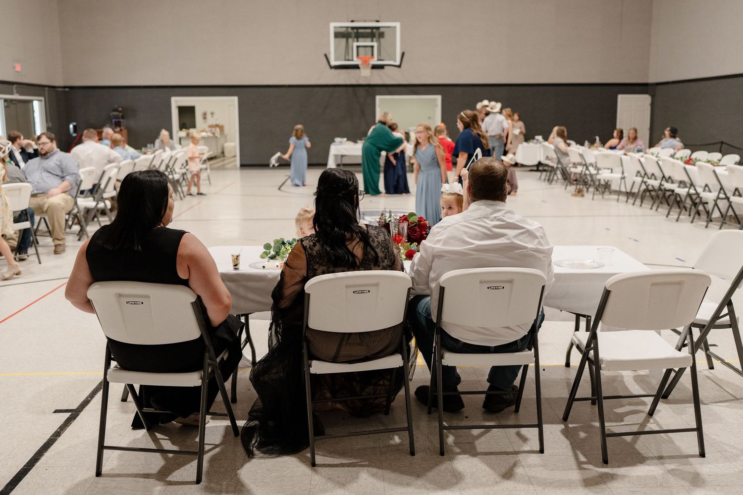 People seated in folding chairs at a large indoor event with rows of white tables and a basketball hoop visible in the background.