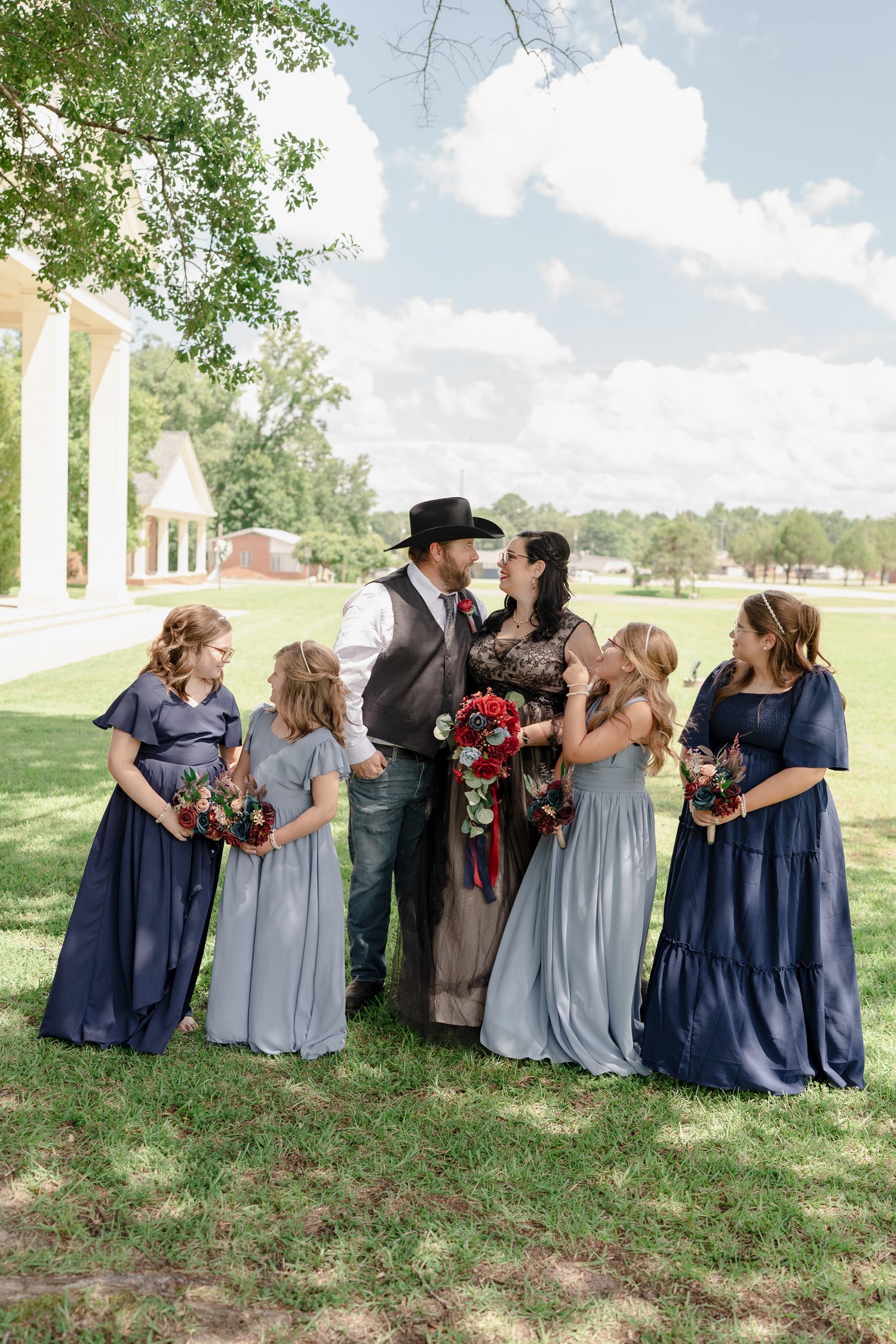 Wedding party in navy and light blue dresses poses playfully on grassy hill with trees in background.