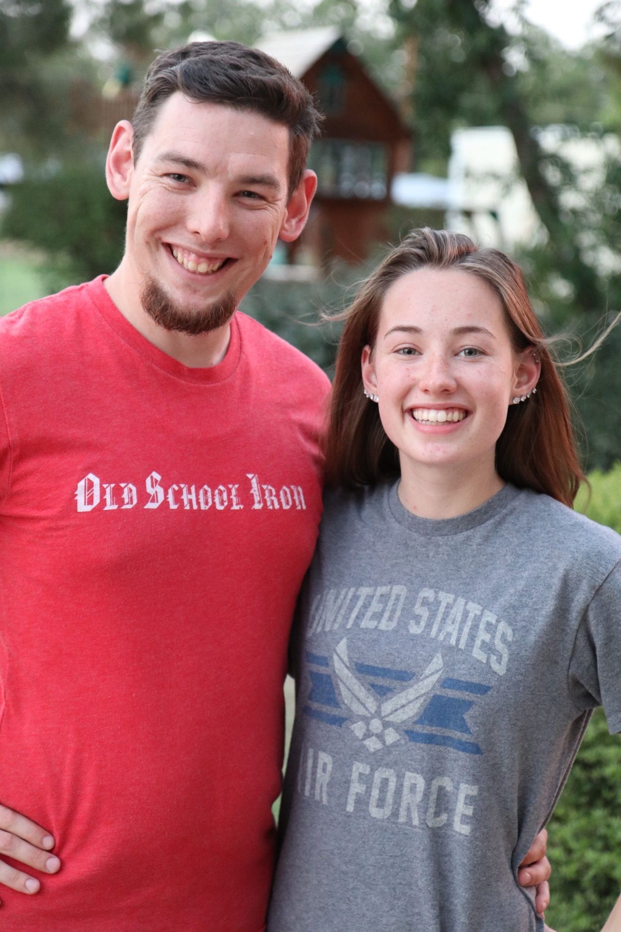 Two people smile together outdoors, wearing red and gray t-shirts.
