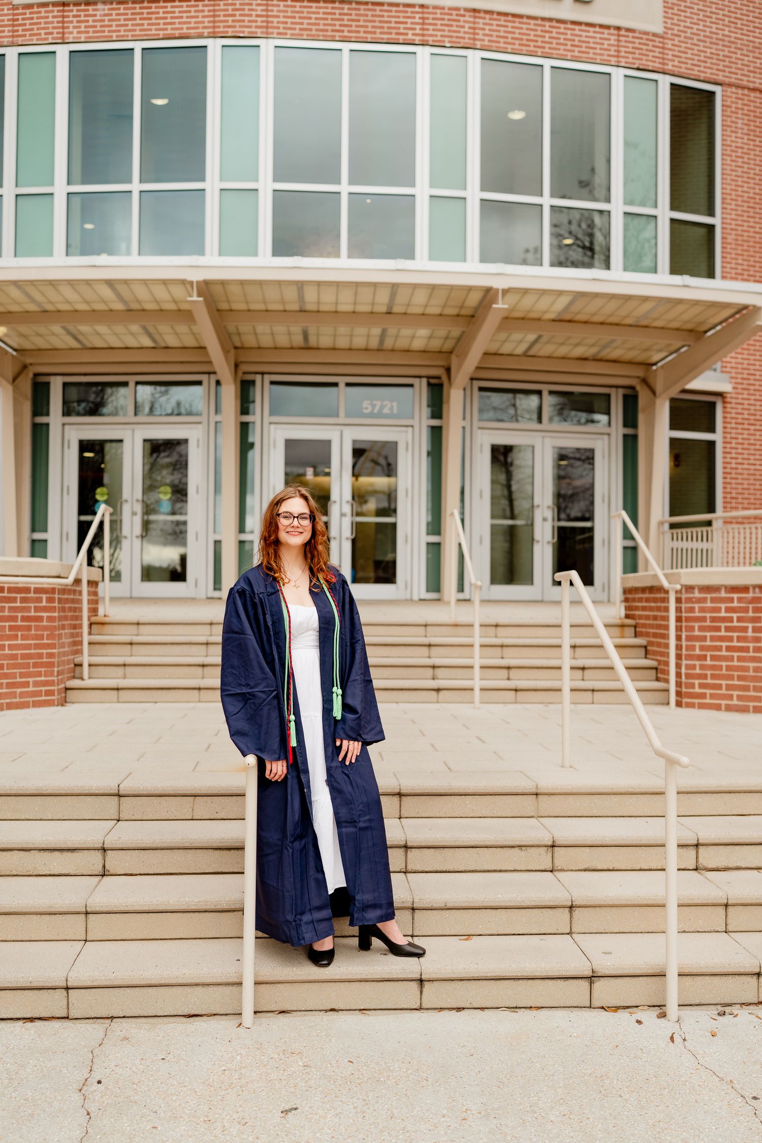 Graduate stands in front of Health Sciences building wearing navy graduation gown and glasses.