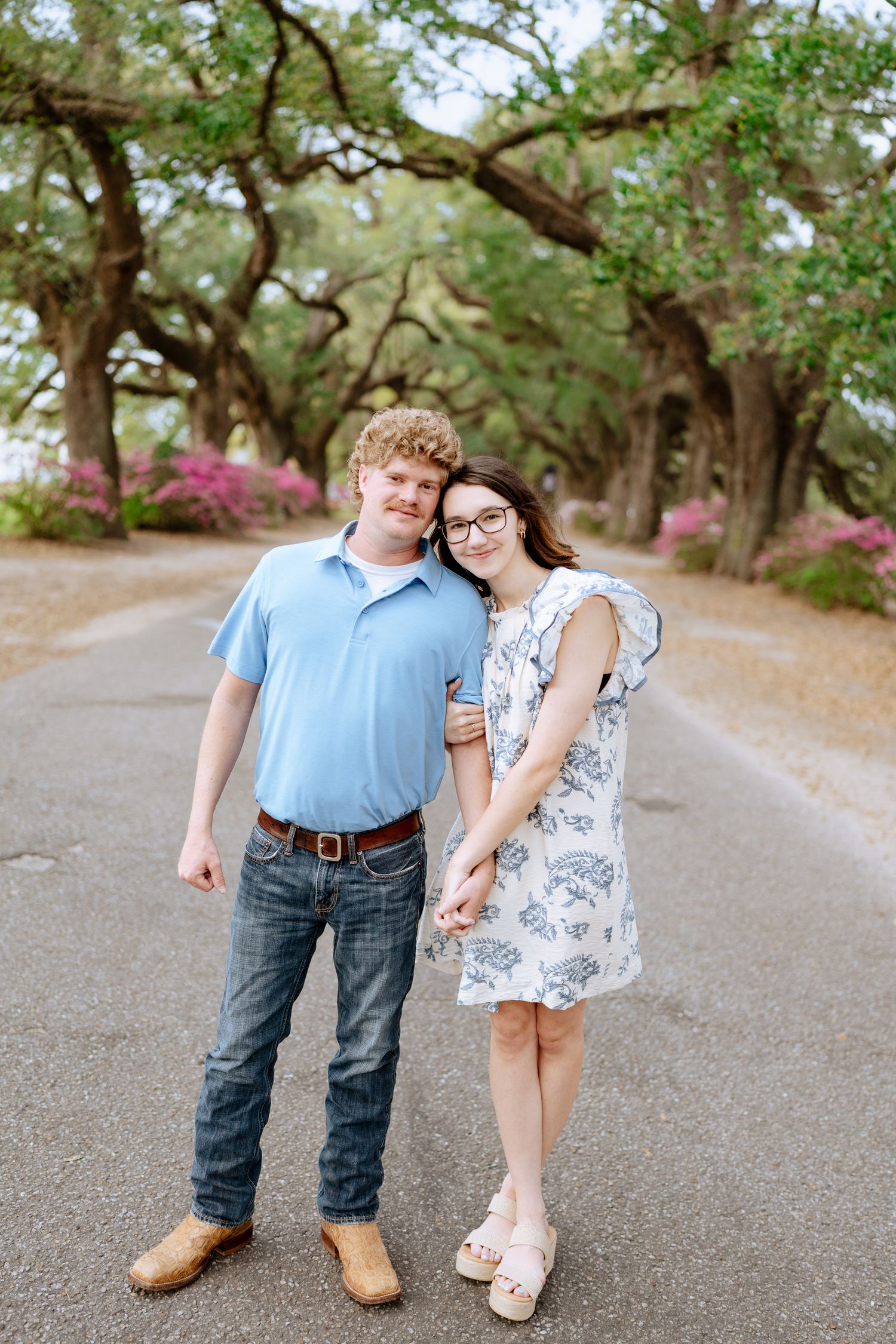 Couple in coordinated outfits share playful moments on path beneath oak trees.