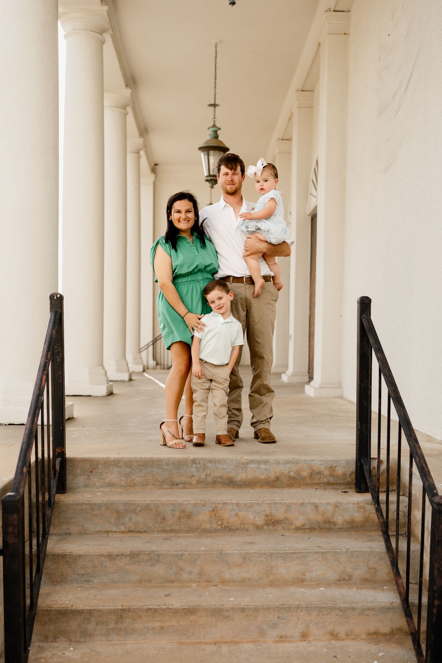 Family of four standing on white columned porch steps with hanging lantern.