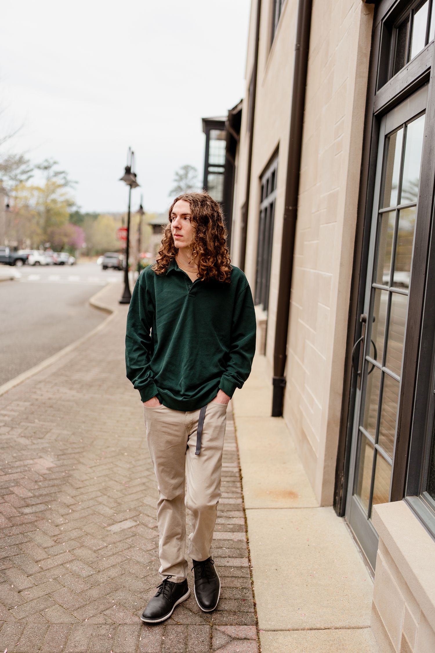 Person in dark green sweatshirt and khaki pants standing on brick sidewalk near storefronts.