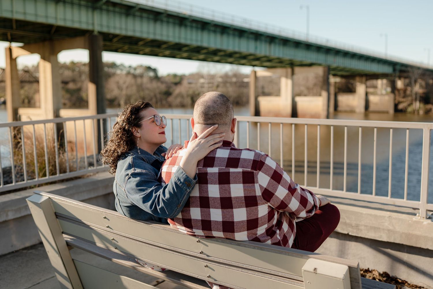 A couple sits together on a wooden bench overlooking a bridge and river during golden hour.