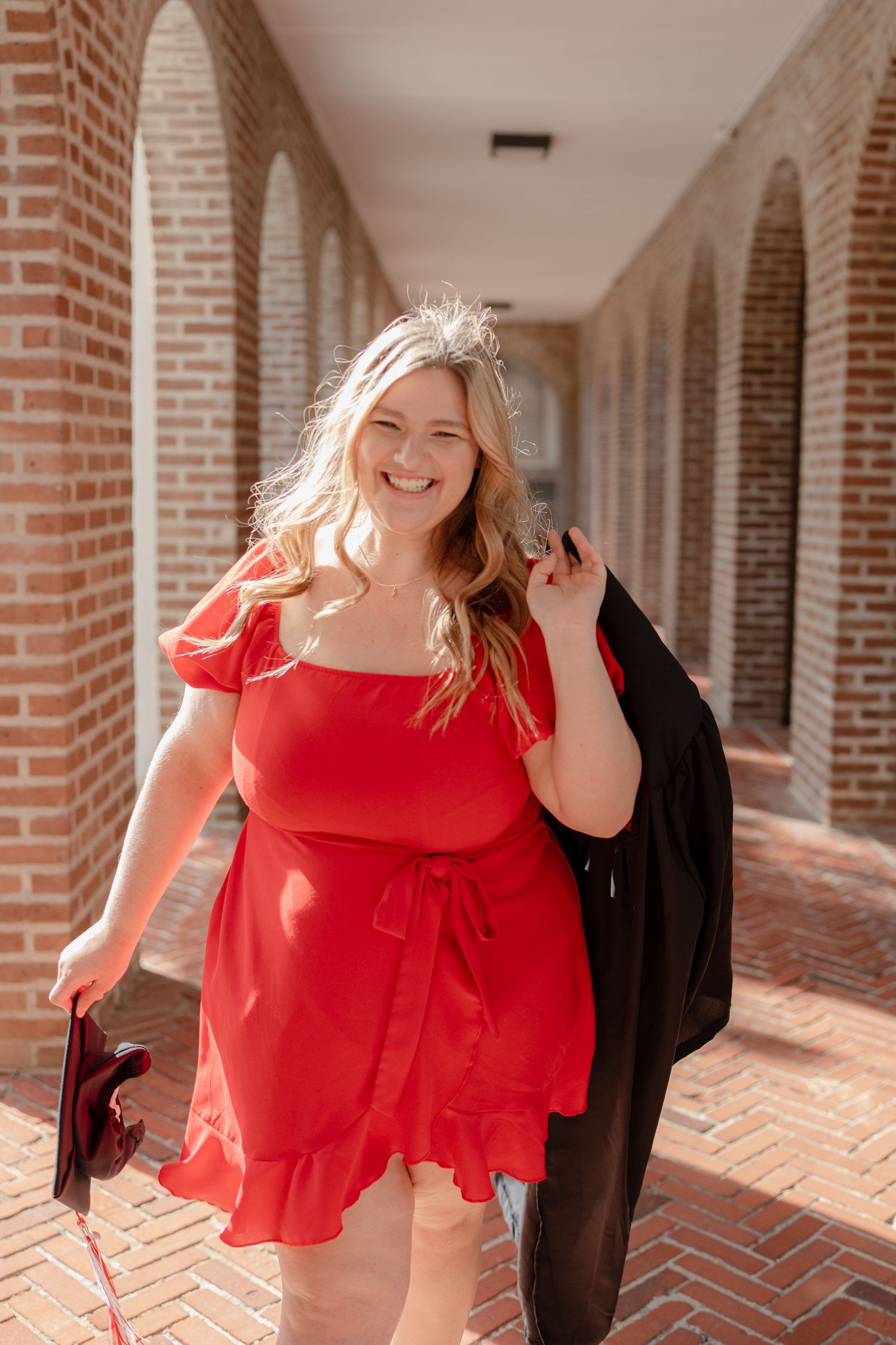 A joyful graduate in a red dress poses in a brick archway corridor while holding graduation regalia and diploma.