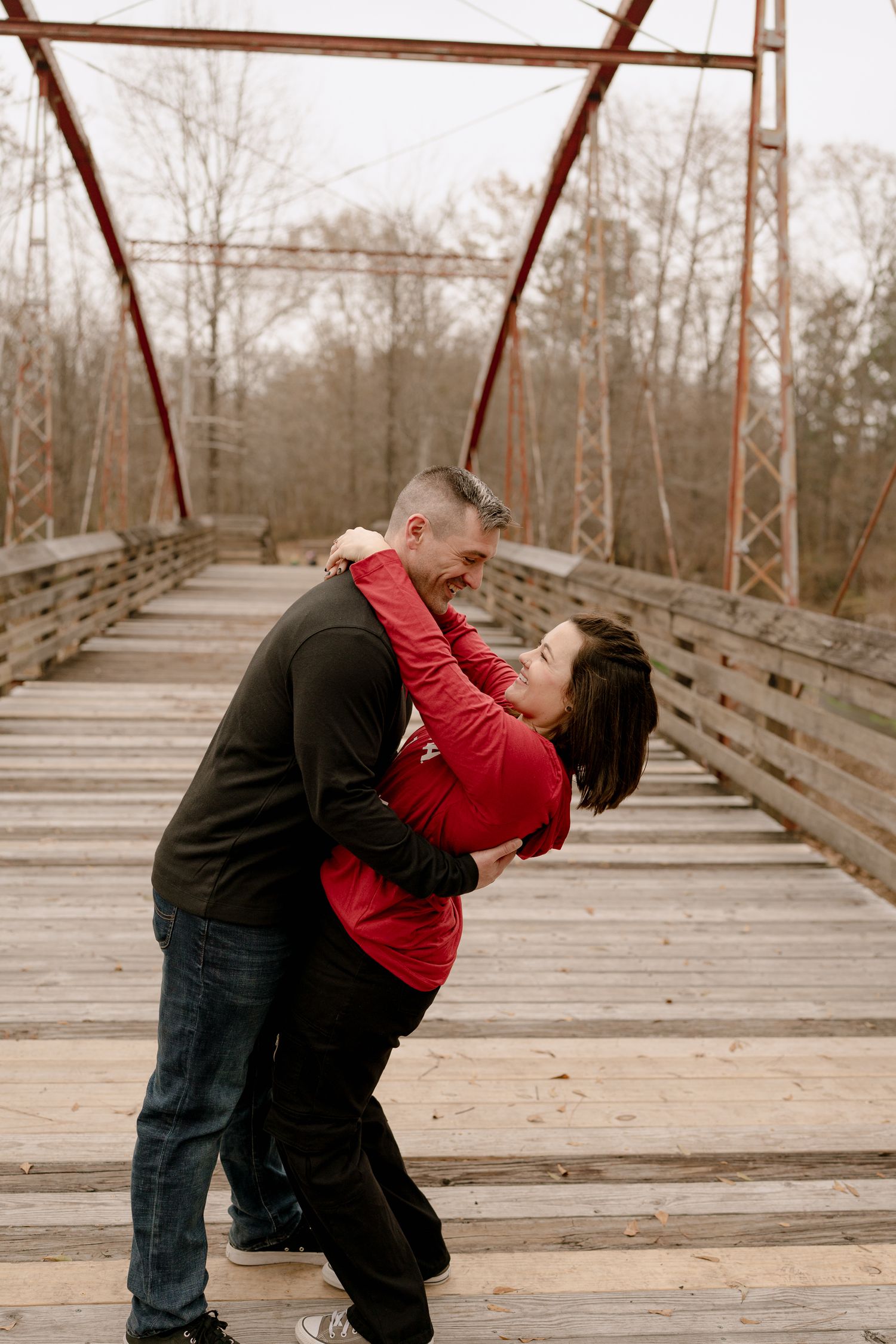 A couple shares a romantic dance pose on a rustic wooden bridge with red metal trusses.