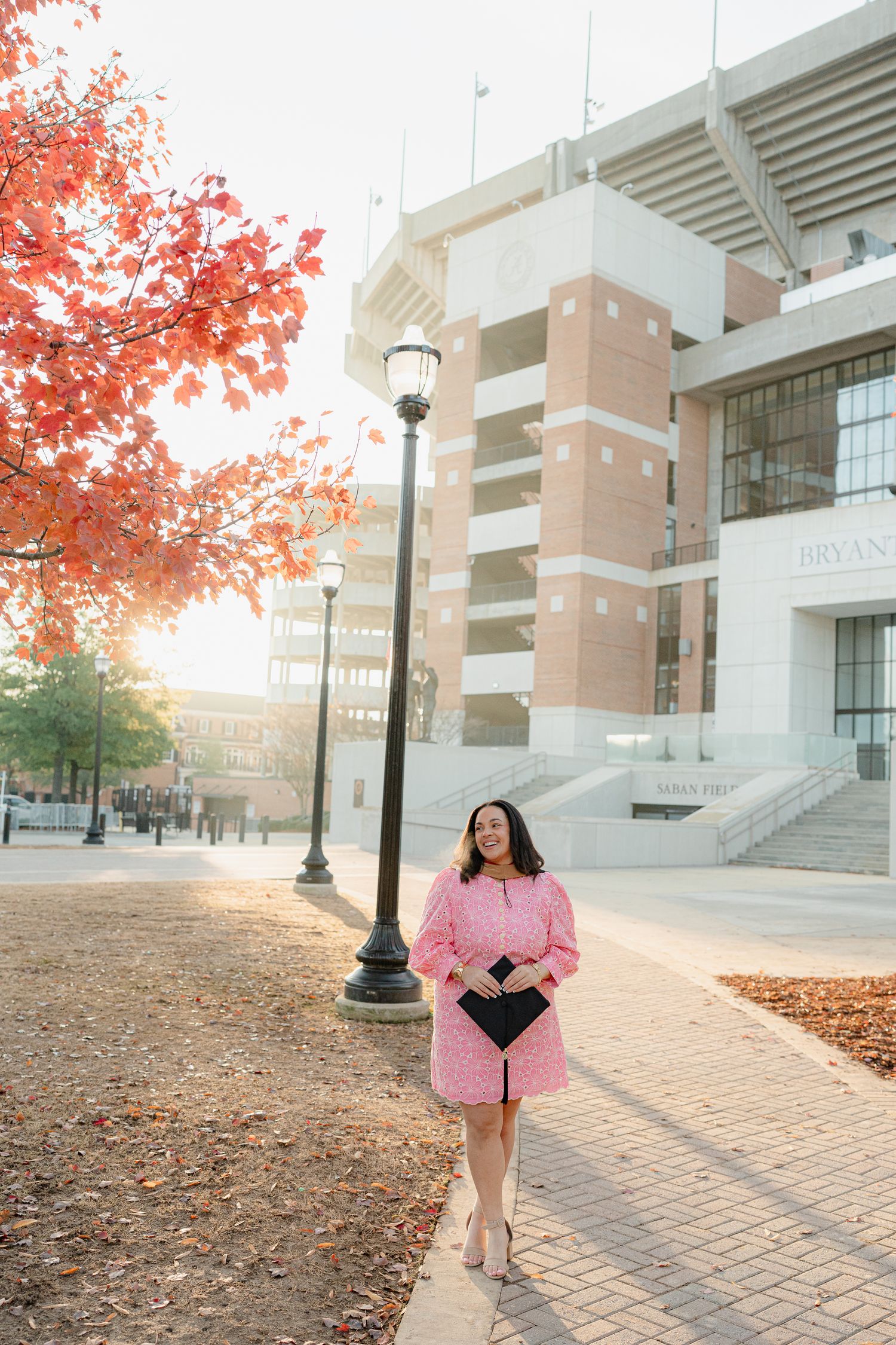 Pink dress and graduation gown against a stadium backdrop with autumn foliage at sunset.