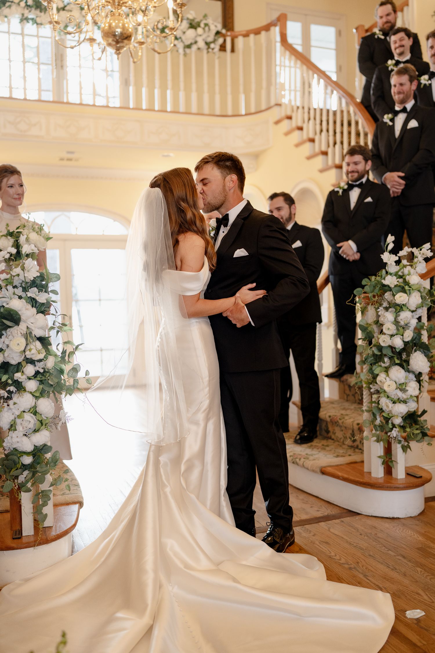 Newlyweds share intimate kiss during ceremony beneath ornate staircase decorated with white floral arrangements.