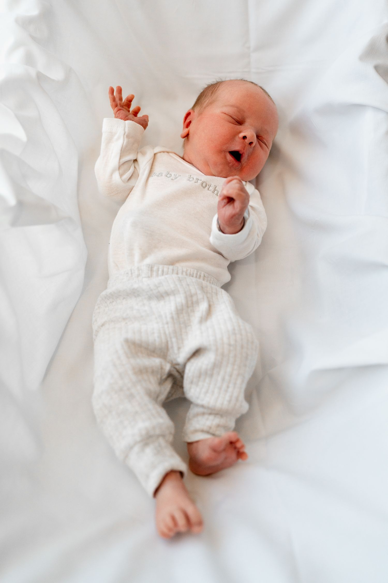Sleeping newborn in white outfit stretching on fresh white linens.