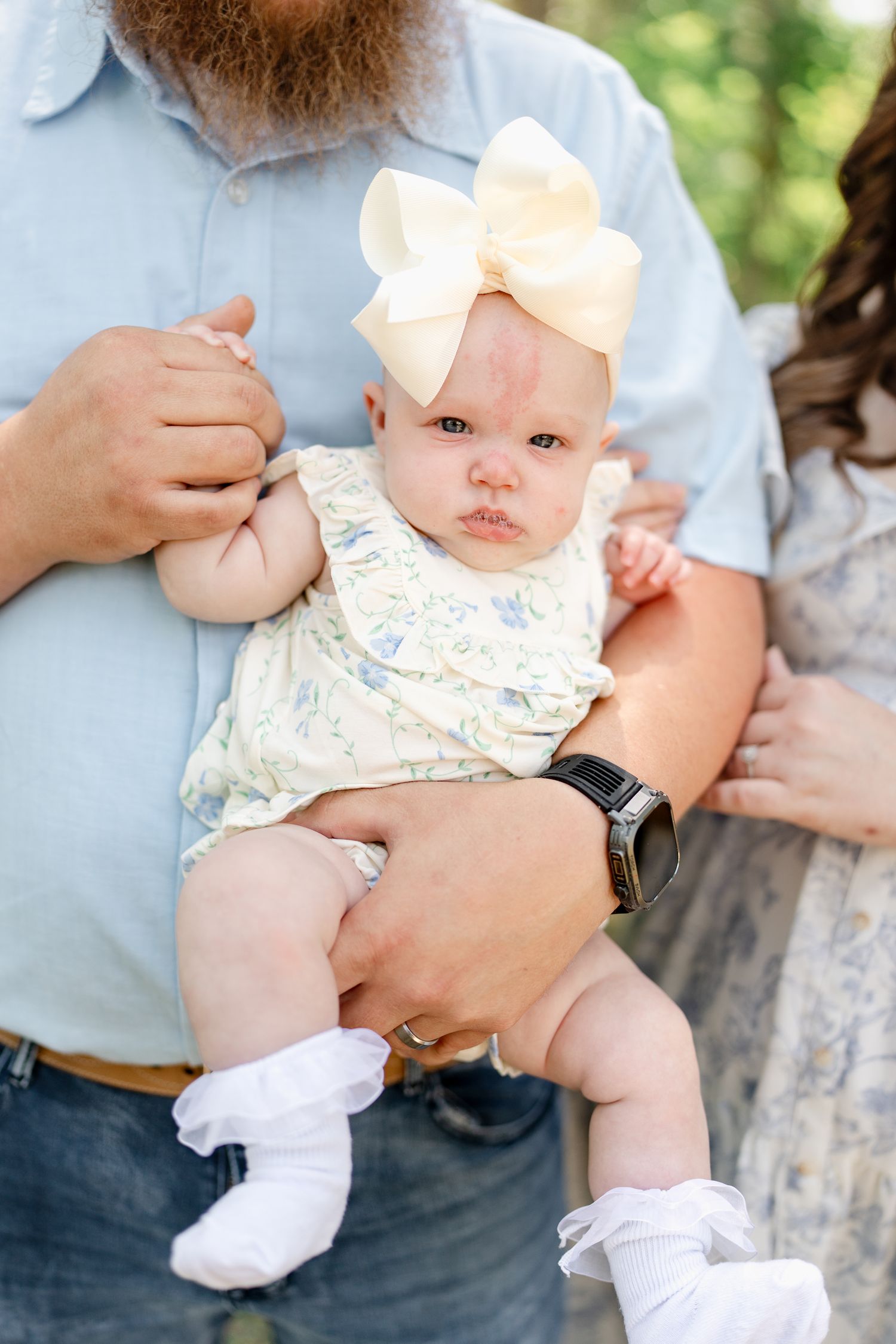 Baby wearing bow and floral outfit held gently in outdoor photo session.