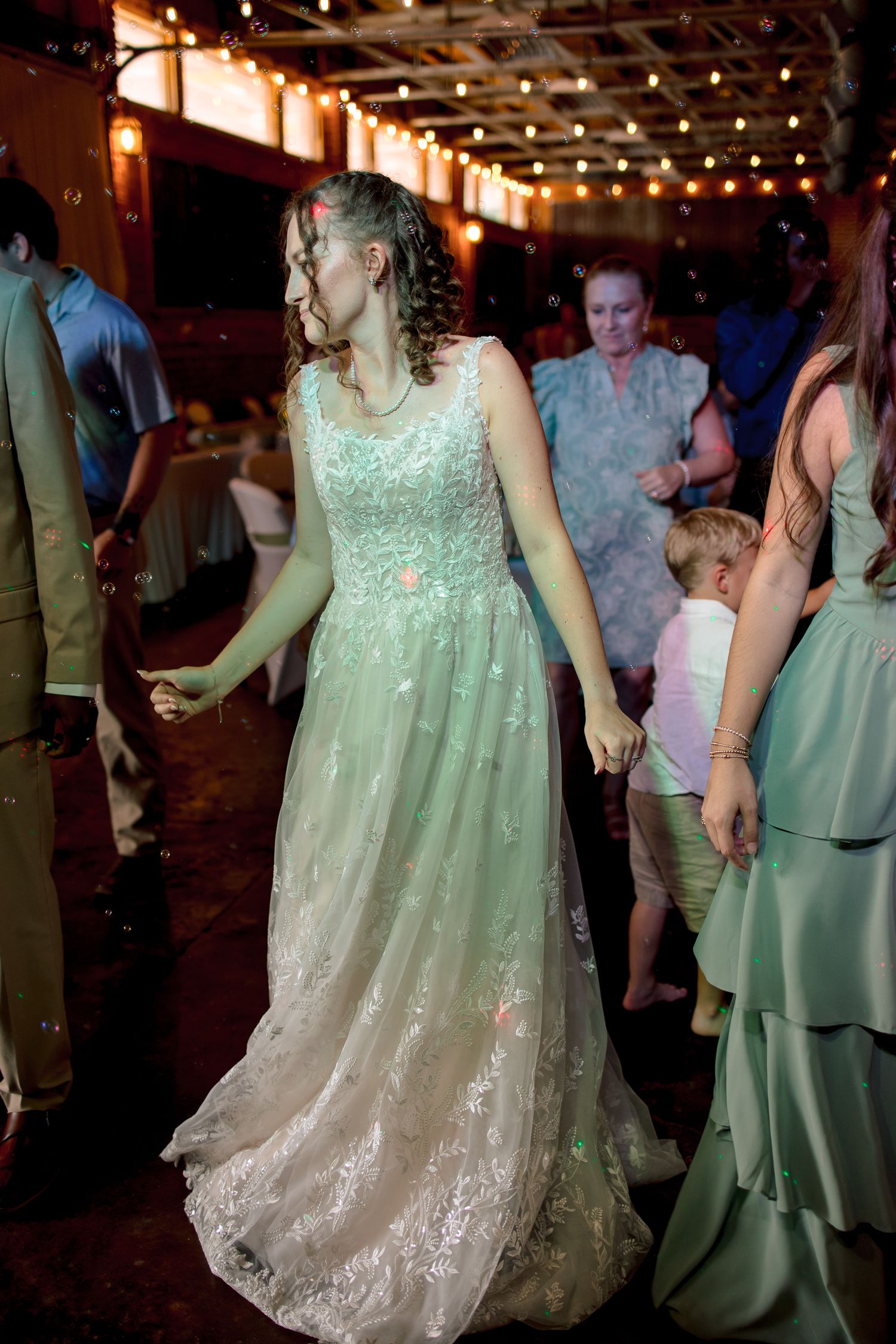 Bride in lace gown dancing with guests under string lights at lively rustic wedding reception venue.