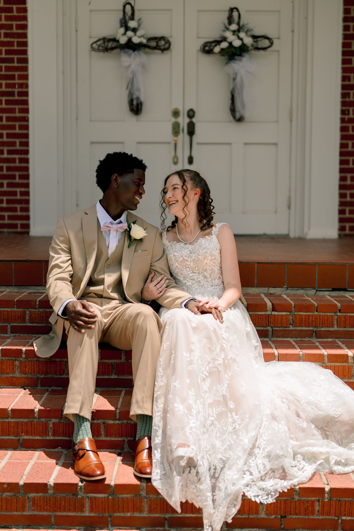 Interracial couple in wedding attire sitting on brick church steps, groom in tan suit, bride in lace gown, smiling at each other.