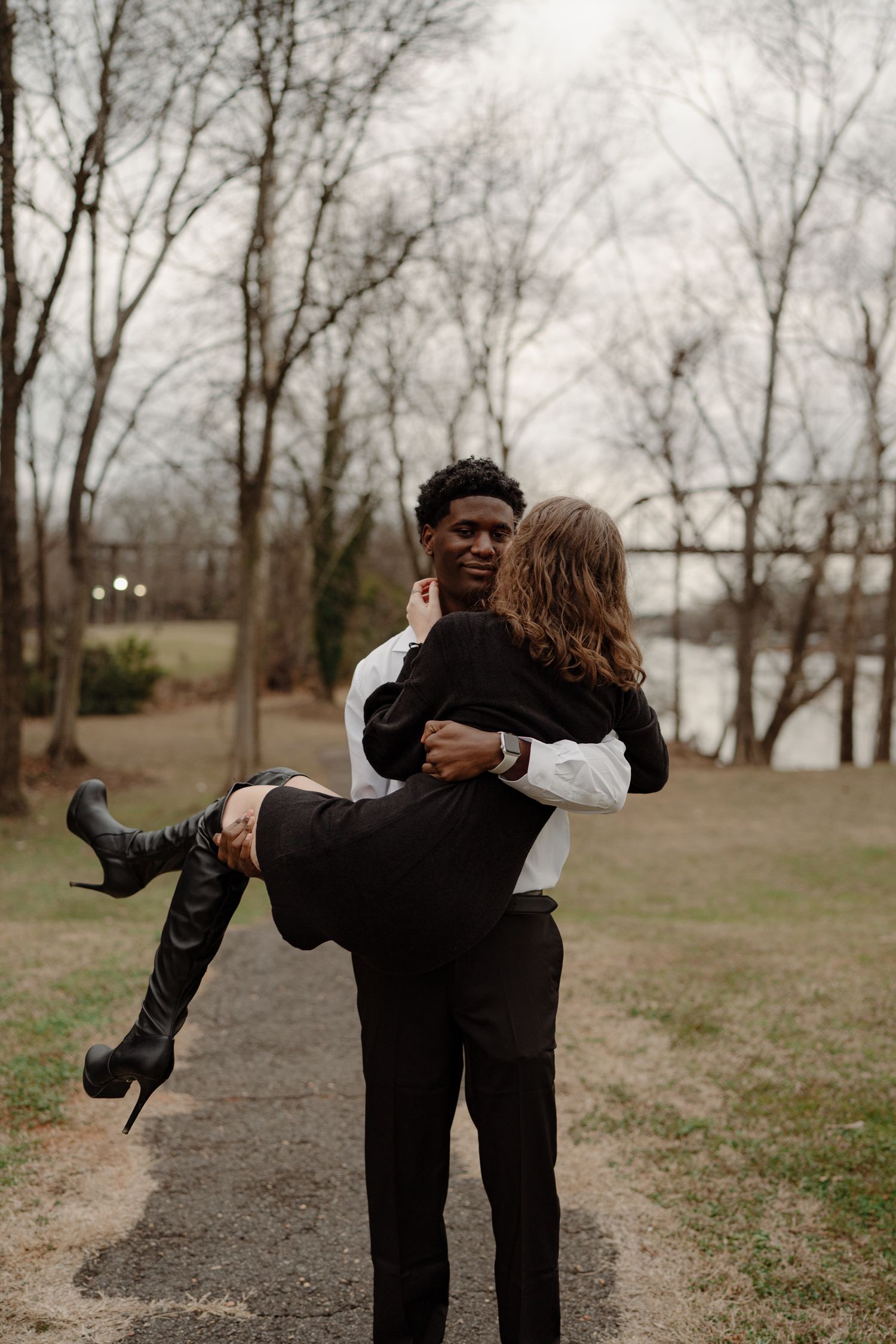 Man in white shirt lifting woman in black dress and boots outdoors near bare winter trees.