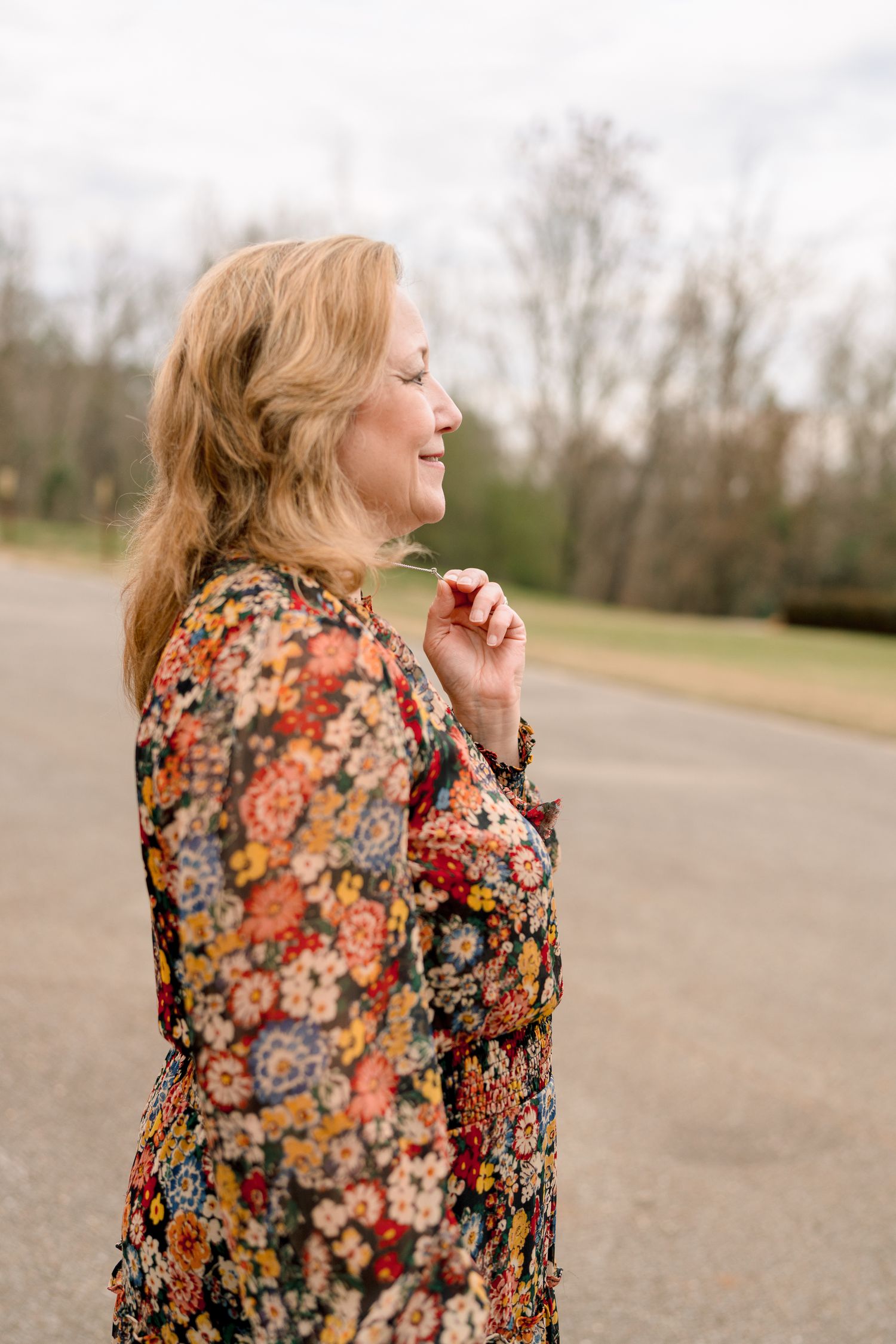 Smiling middle-aged woman in a floral dress standing outdoors on a road with trees behind her.