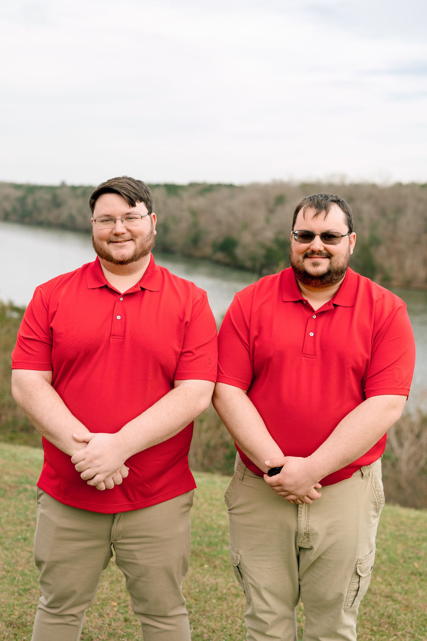 Two brothers in matching red polo shirts smile together outdoors by a river.