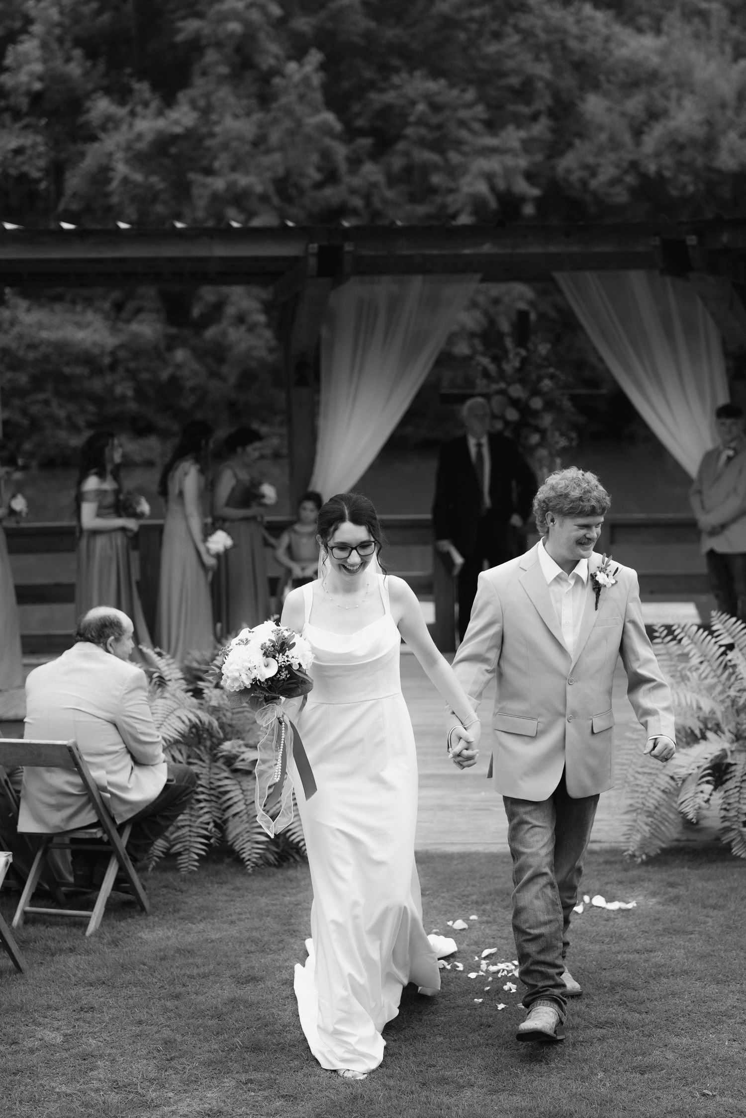Black and white photo of bride and groom walking back down aisle after outdoor barn wedding ceremony.