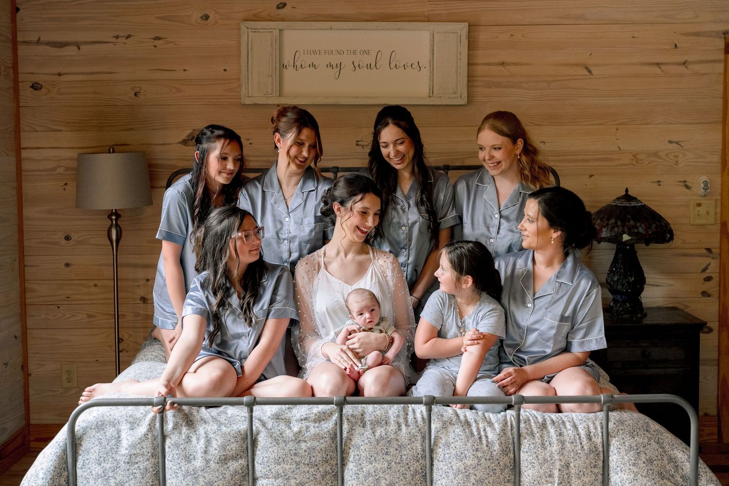 Bride holding newborn baby surrounded by bridesmaids in gray robes on a rustic bed before the wedding.