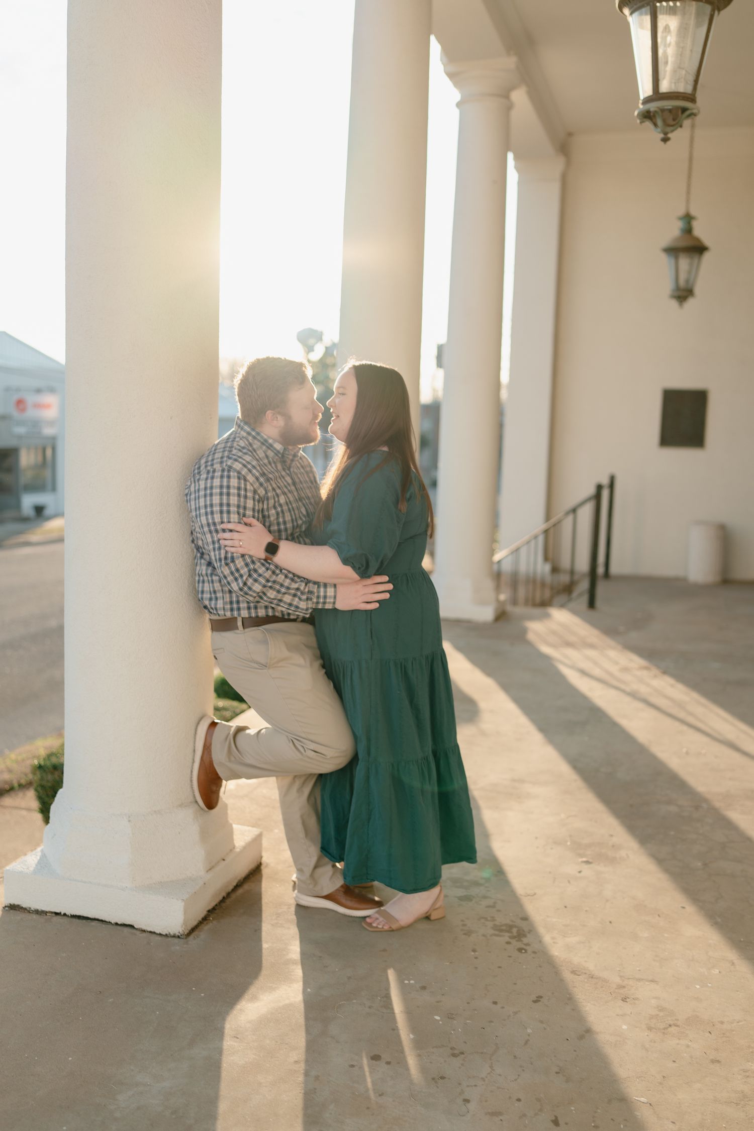 Couple sharing a romantic almost-kiss leaning against white column at golden hour in elegant outdoor venue.