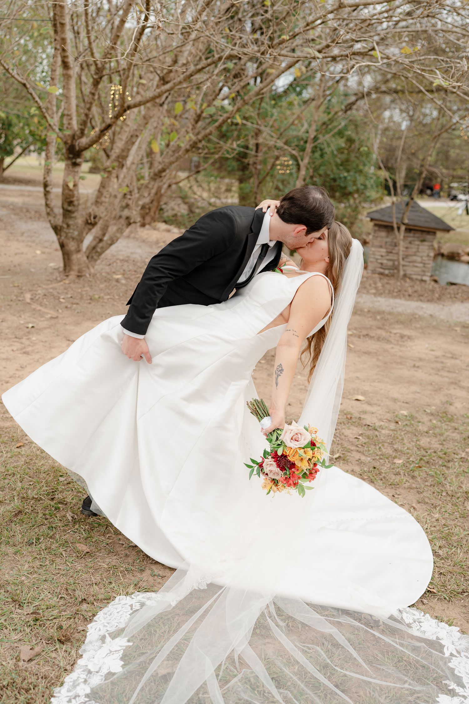 Romantic wedding couple in white dress and black suit dips and kisses in an outdoor wooded setting.
