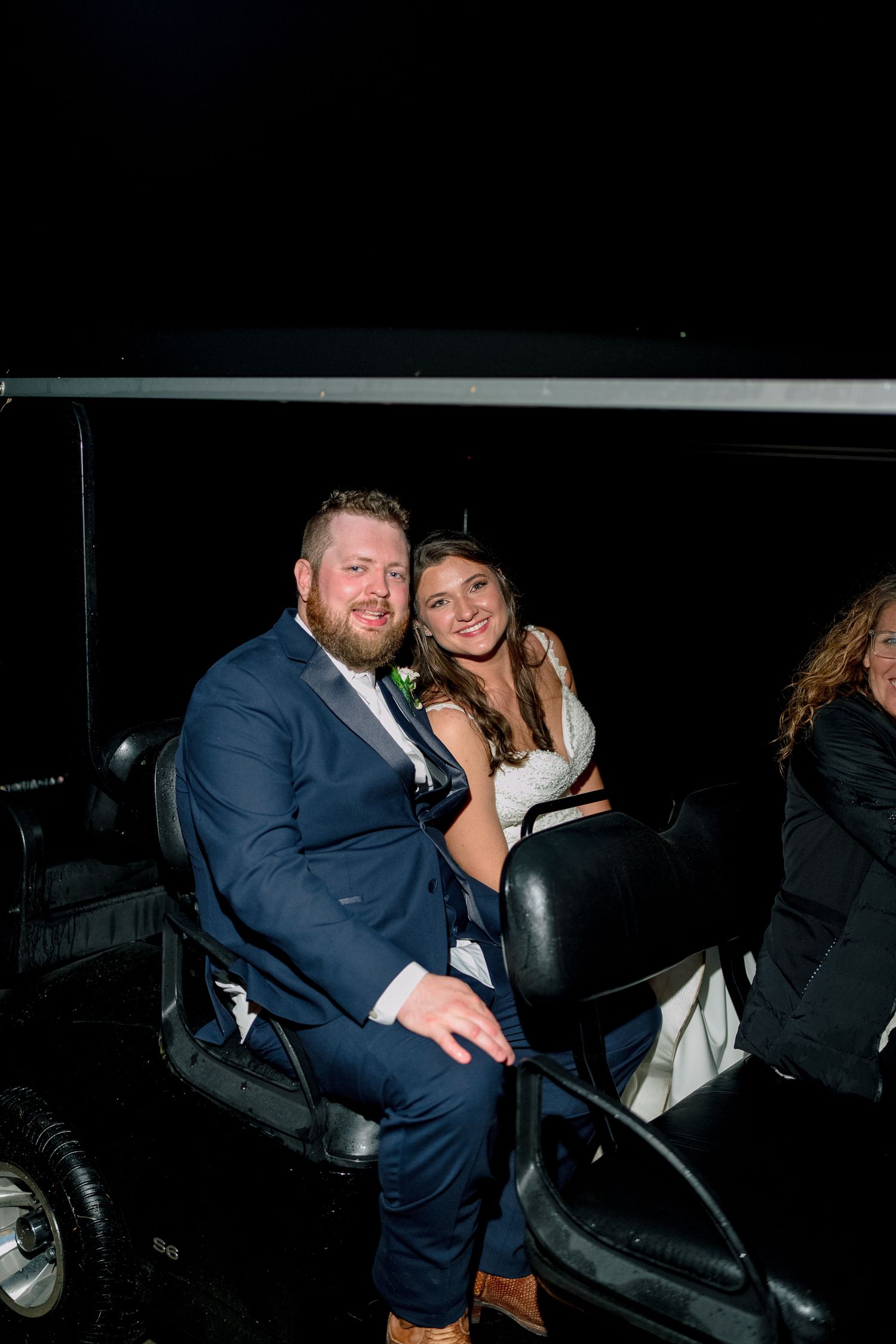Bride and groom smile together while riding a golf cart at night after their wedding reception.