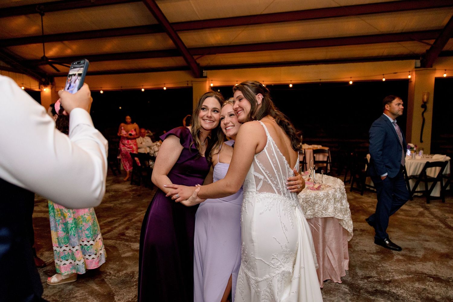 Bride in backless gown hugging guests while being photographed at wedding reception.