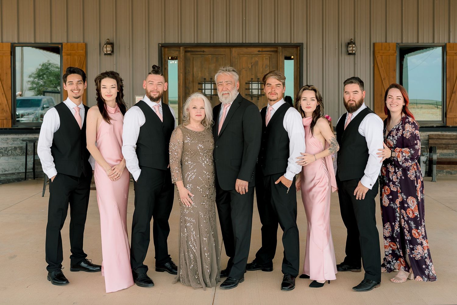 Large family group poses in formal wedding attire outside elegant barn venue on overcast day.