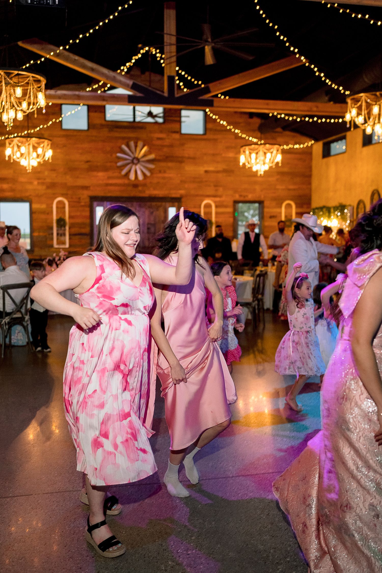 Wedding guests in pink and floral dresses dance joyfully under string lights and chandeliers in a rustic barn reception venue.