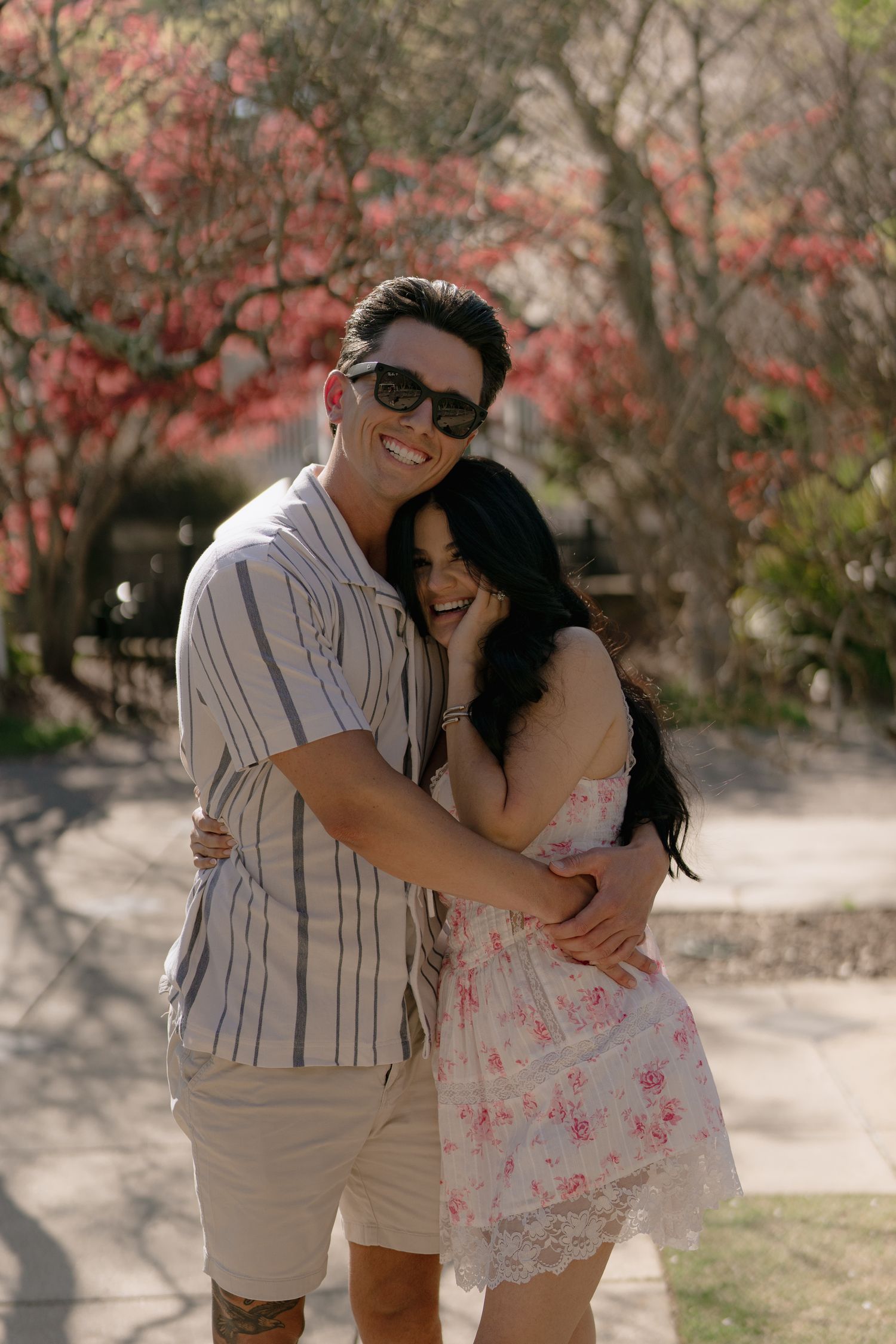 Couple embracing and smiling outdoors near pink flowering trees on a sunny day.