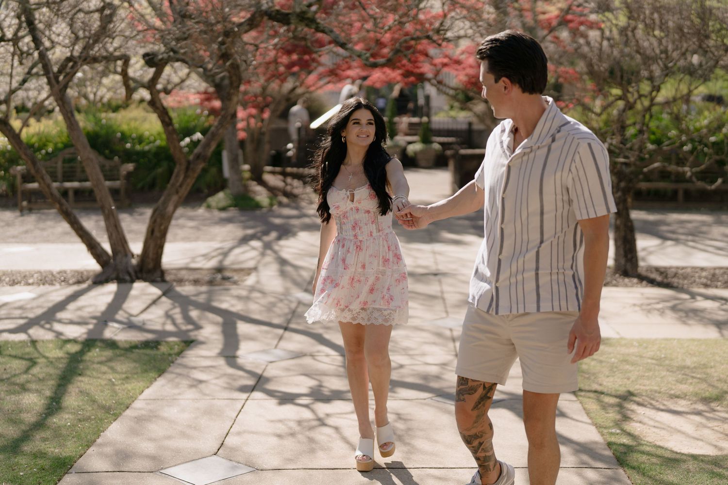 Couple holding hands walking on a sunny garden path surrounded by blooming trees in a romantic outdoor setting.