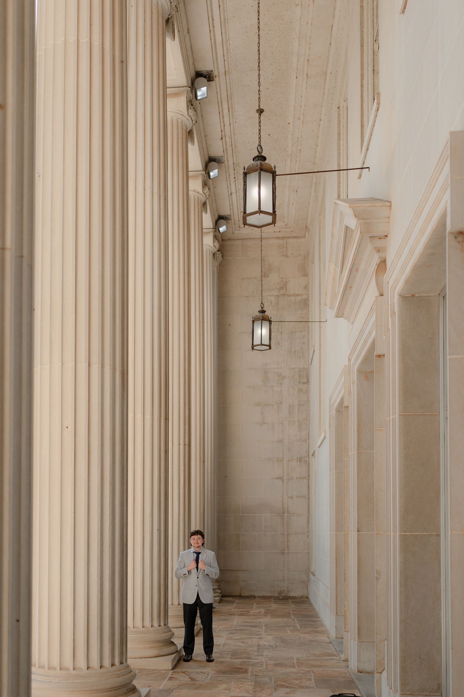 Young man in gray suit stands between towering classical columns with hanging lanterns in grand hallway.