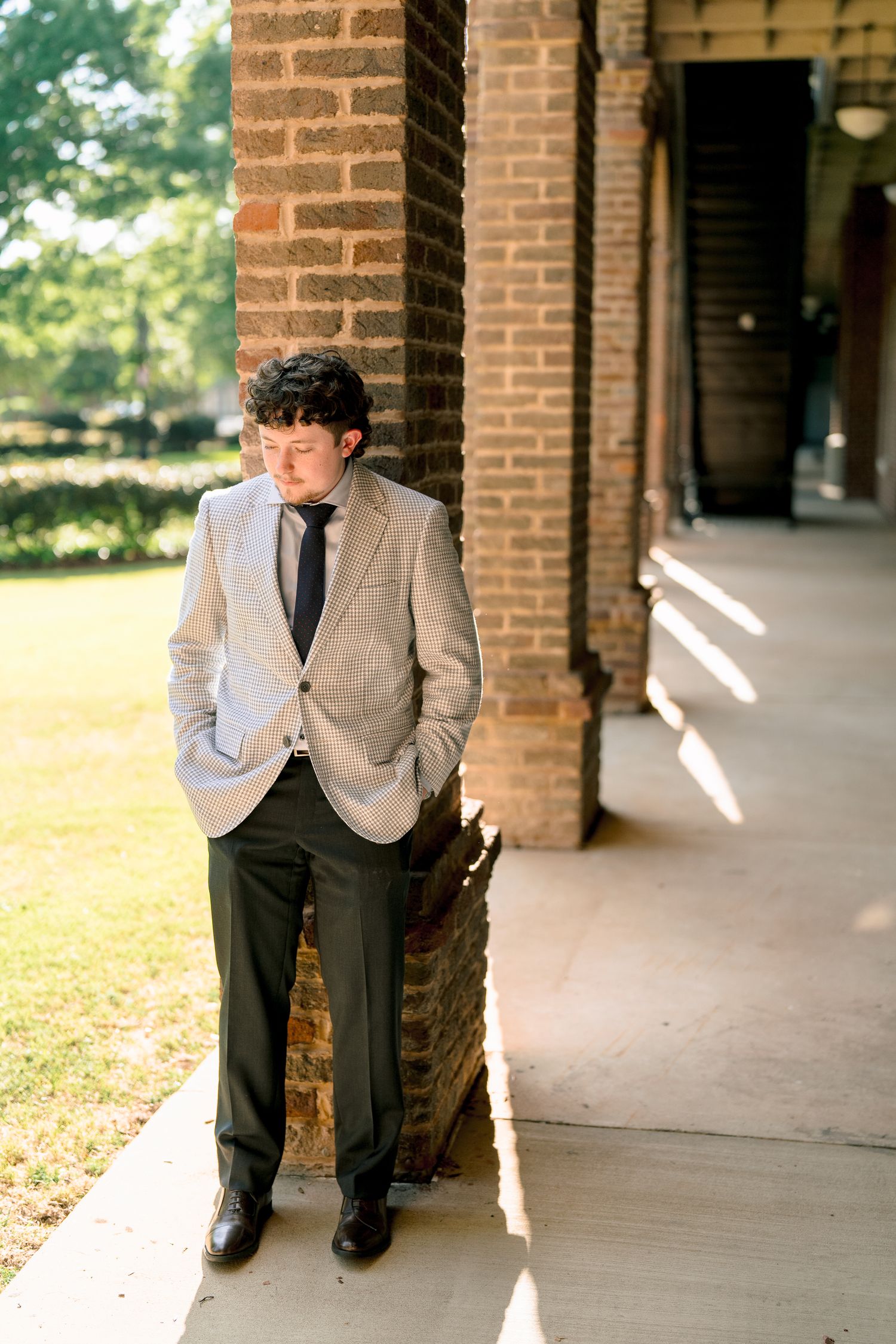 Young man in gray blazer with hands in pockets leans against brick column looking down in warm sunlight.