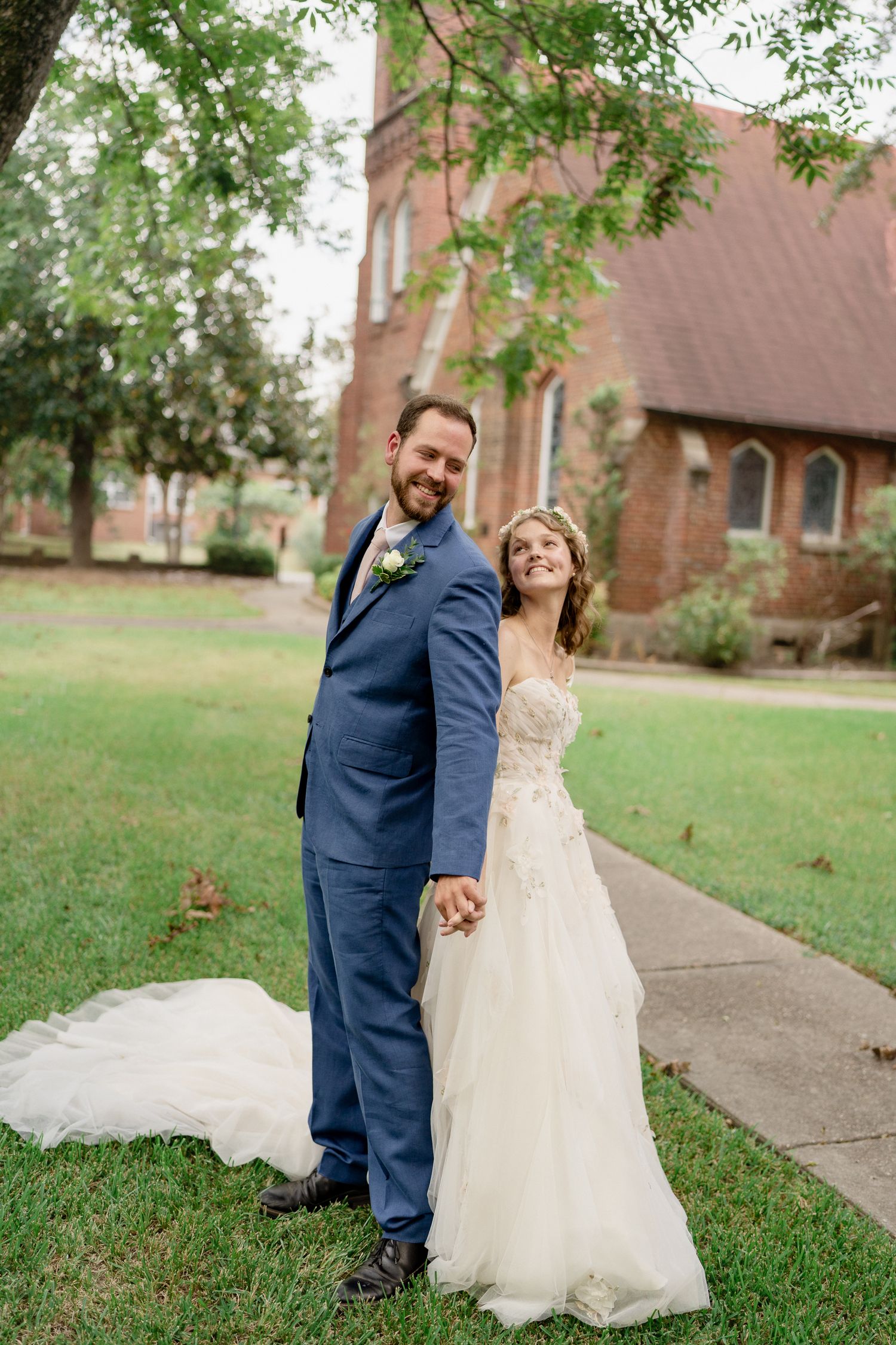 A couple in wedding attire share joyful moments together on a grassy lawn with a brick church in the background.