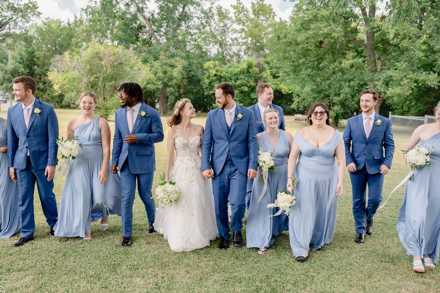Bridal party sits in casual formation on grass wearing light blue dresses and navy suits.