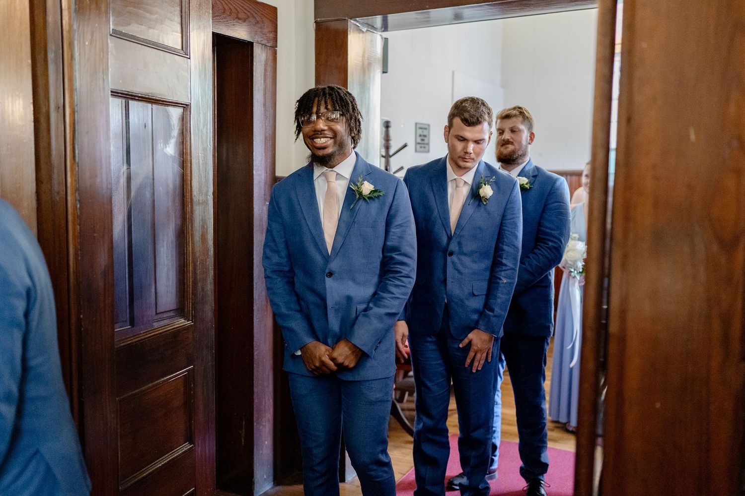 A wedding party wearing matching navy blue suits stands together in a wood-paneled hallway.