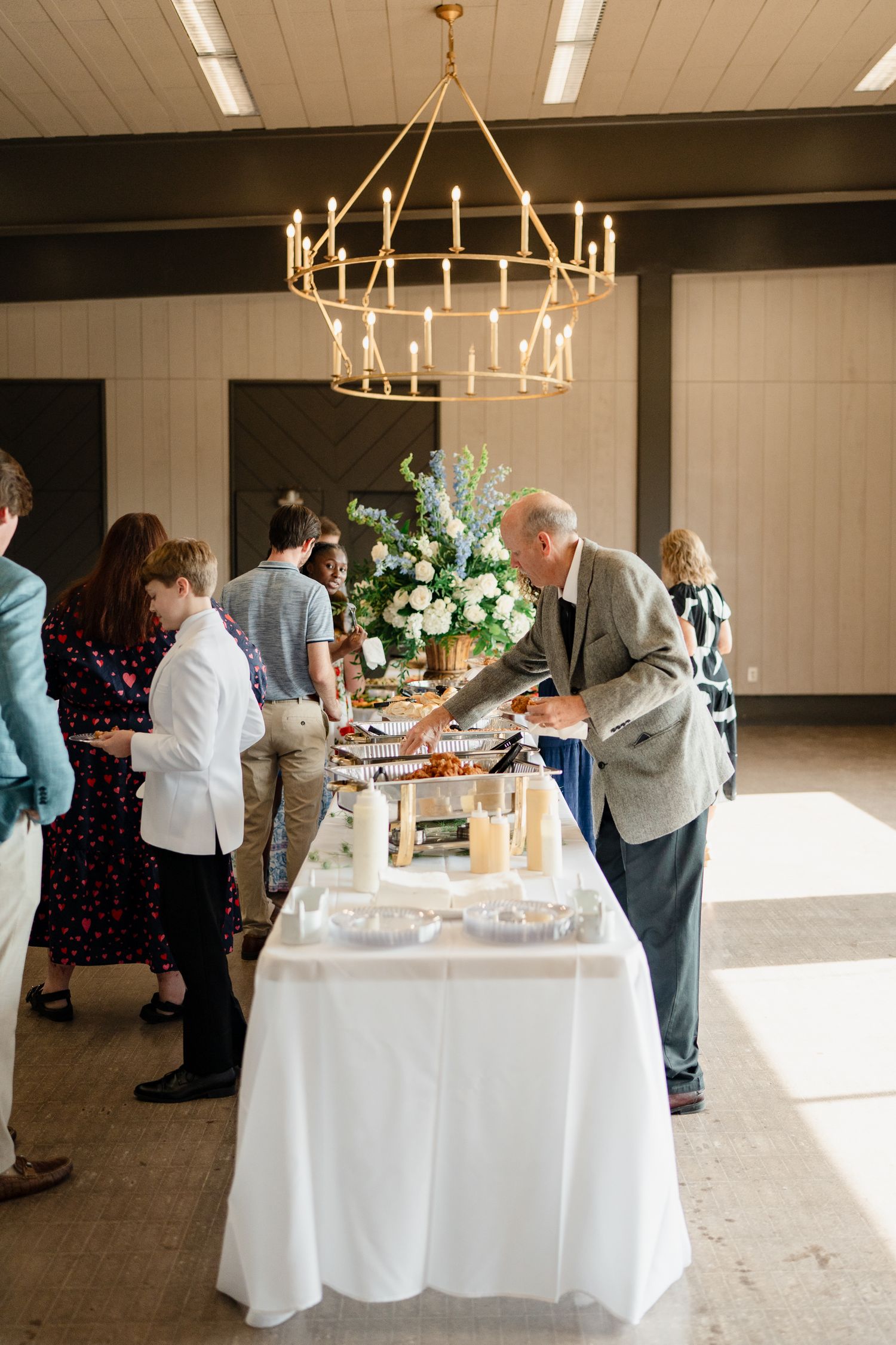 Guests serve themselves at an elegant buffet table with white linens and floral arrangements under modern chandeliers.